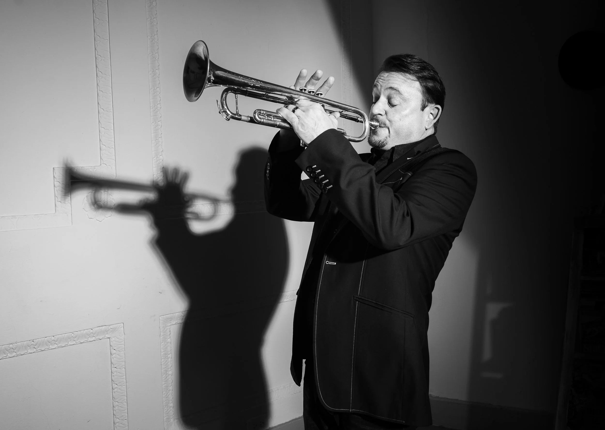 A man in a suit is playing a trumpet with his eyes closed. The image is in black and white, and he is standing against a wall with decorative molding.
