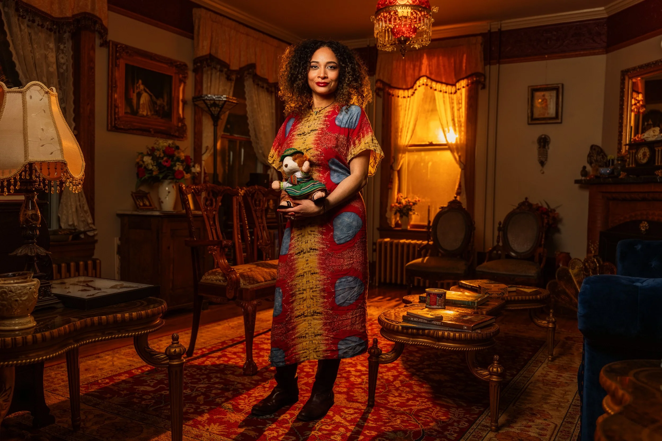 A woman with curly hair wearing a colorful dress holds a small mouse doll in a richly decorated vintage living room with ornate furniture, framed paintings, and warm lighting.