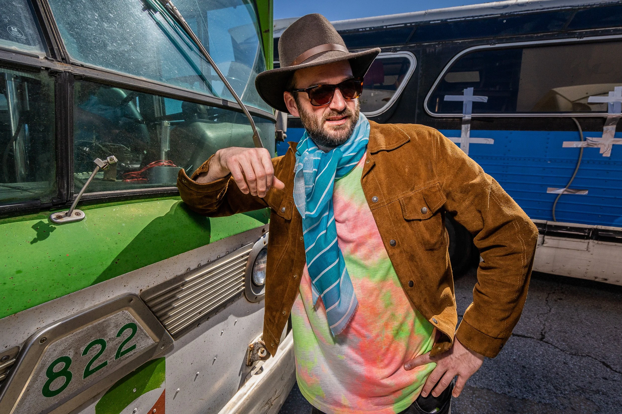 A man wearing a brown jacket, colorful tie-dye shirt, large brown hat, and sunglasses standing beside a green vehicle with a blue bus in the background.