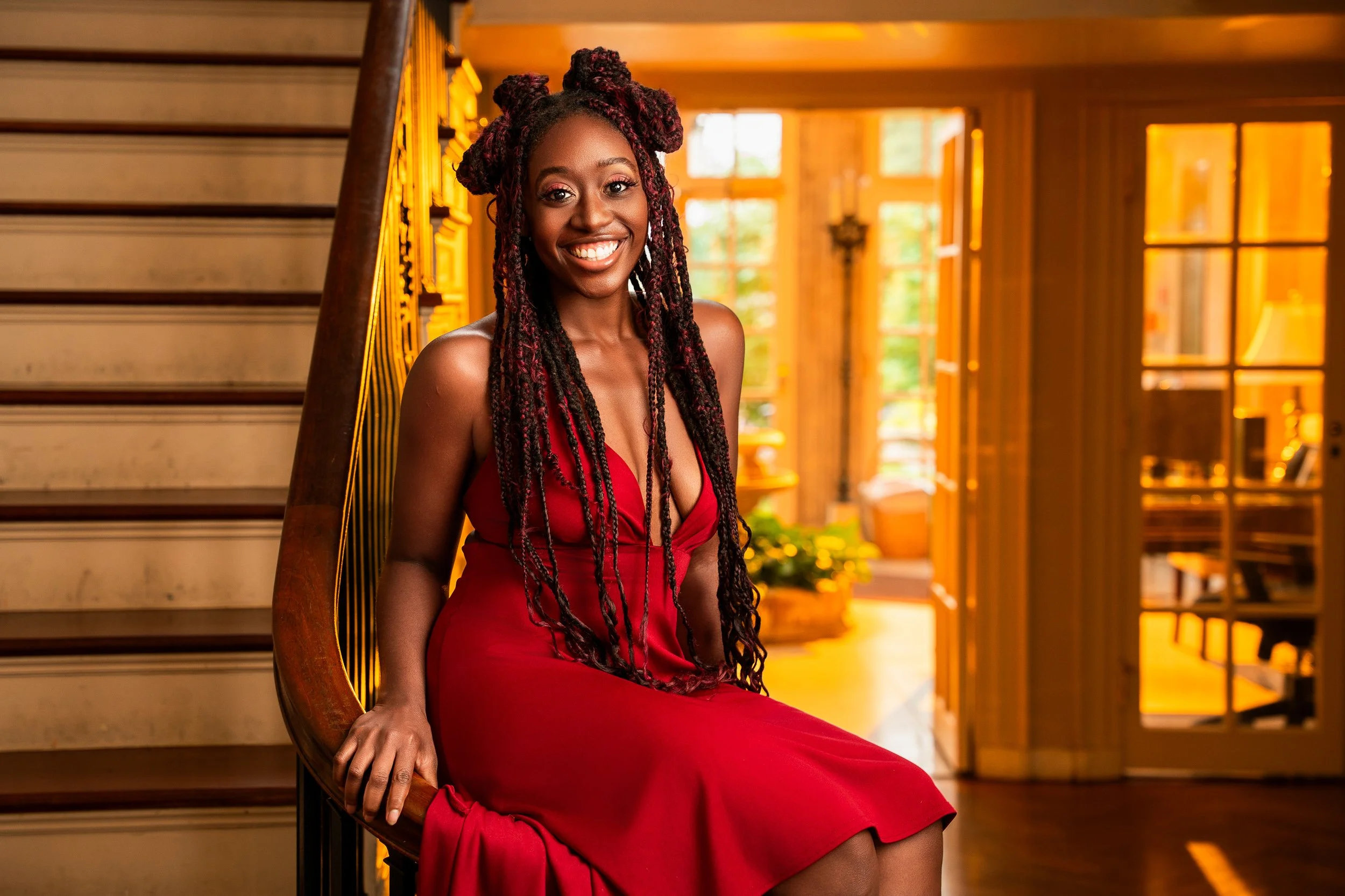 A woman with braided hair and a red dress sitting on a wooden chair in a warmly lit room with large windows and a staircase.