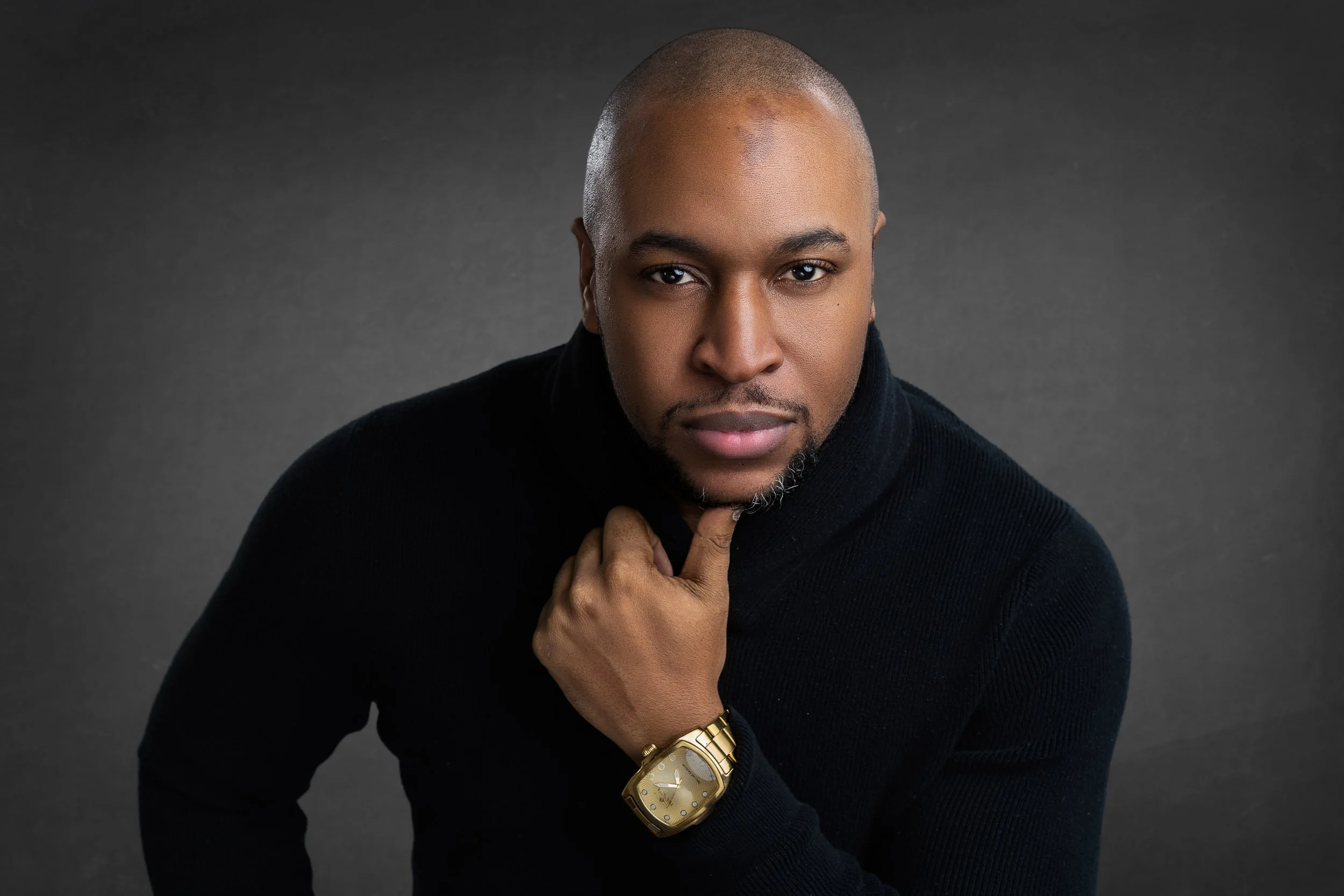 Portrait of a confident Black man with a shaved head, wearing a black sweater, a gold watch, and posing with his hand near his chin against a dark gray background.