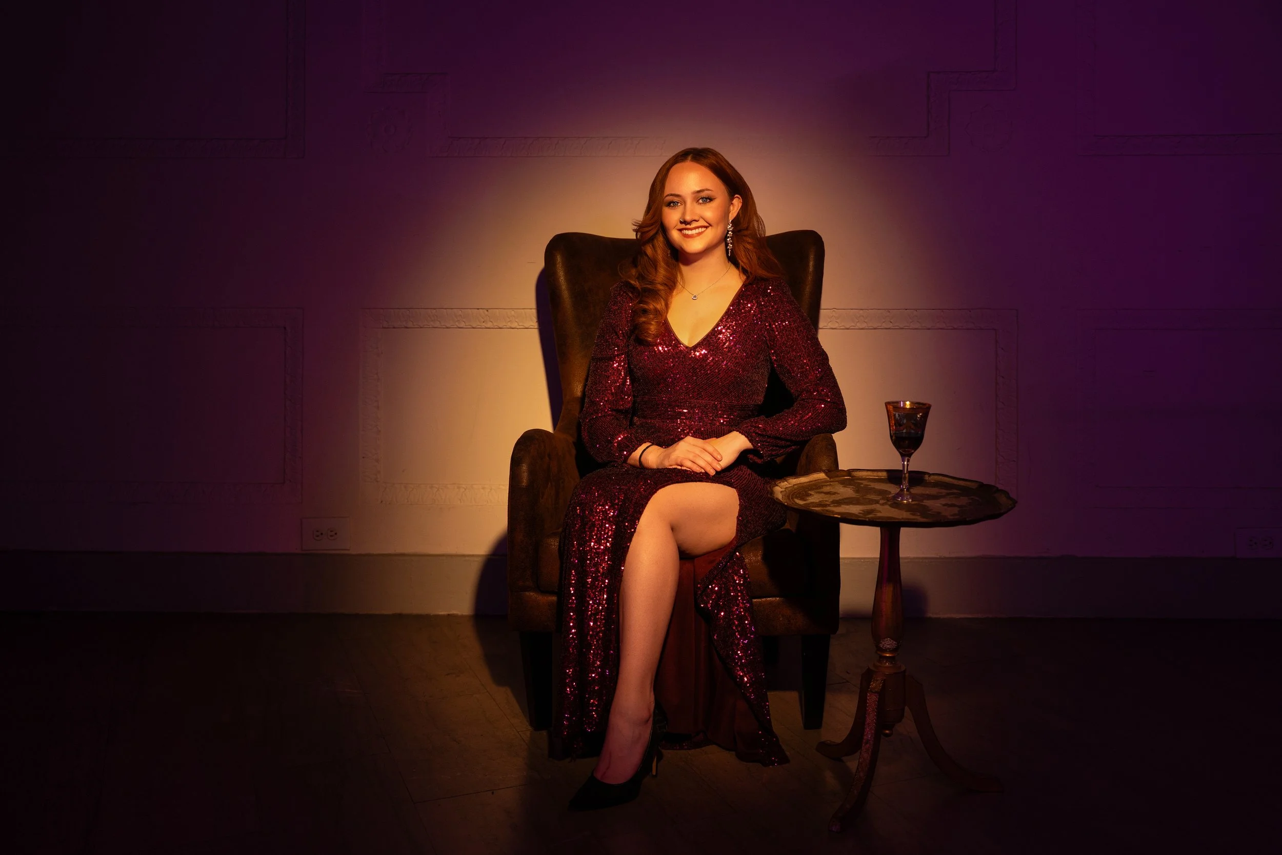 A young woman in a sparkly red dress sitting on a dark armchair with a smile, nightstand with a wine glass beside her, dim lighting creating a cozy atmosphere.