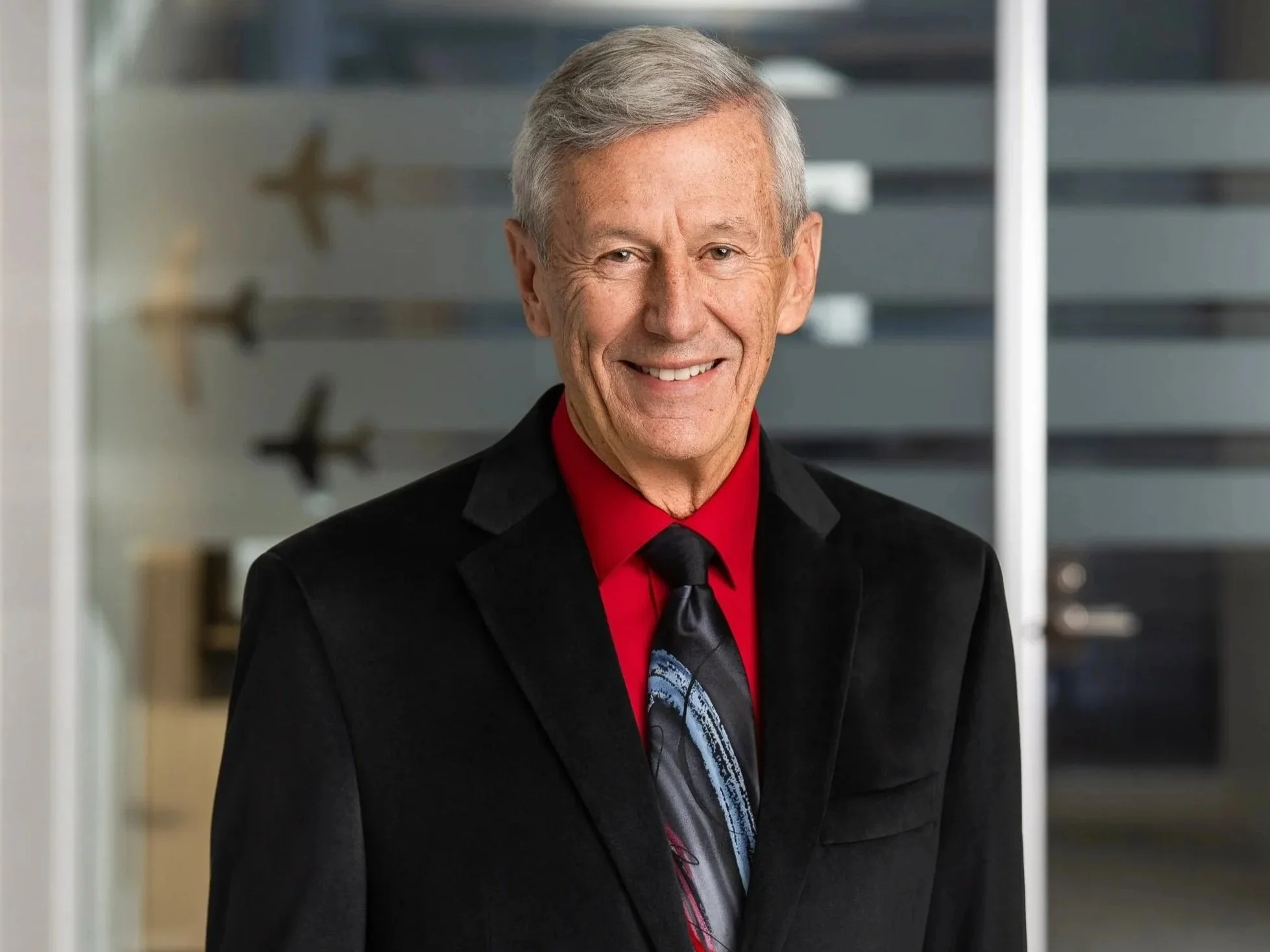 Portrait of an elderly man with gray hair smiling, wearing a black blazer, red shirt, and black tie, standing indoors near a window with a blurred background.