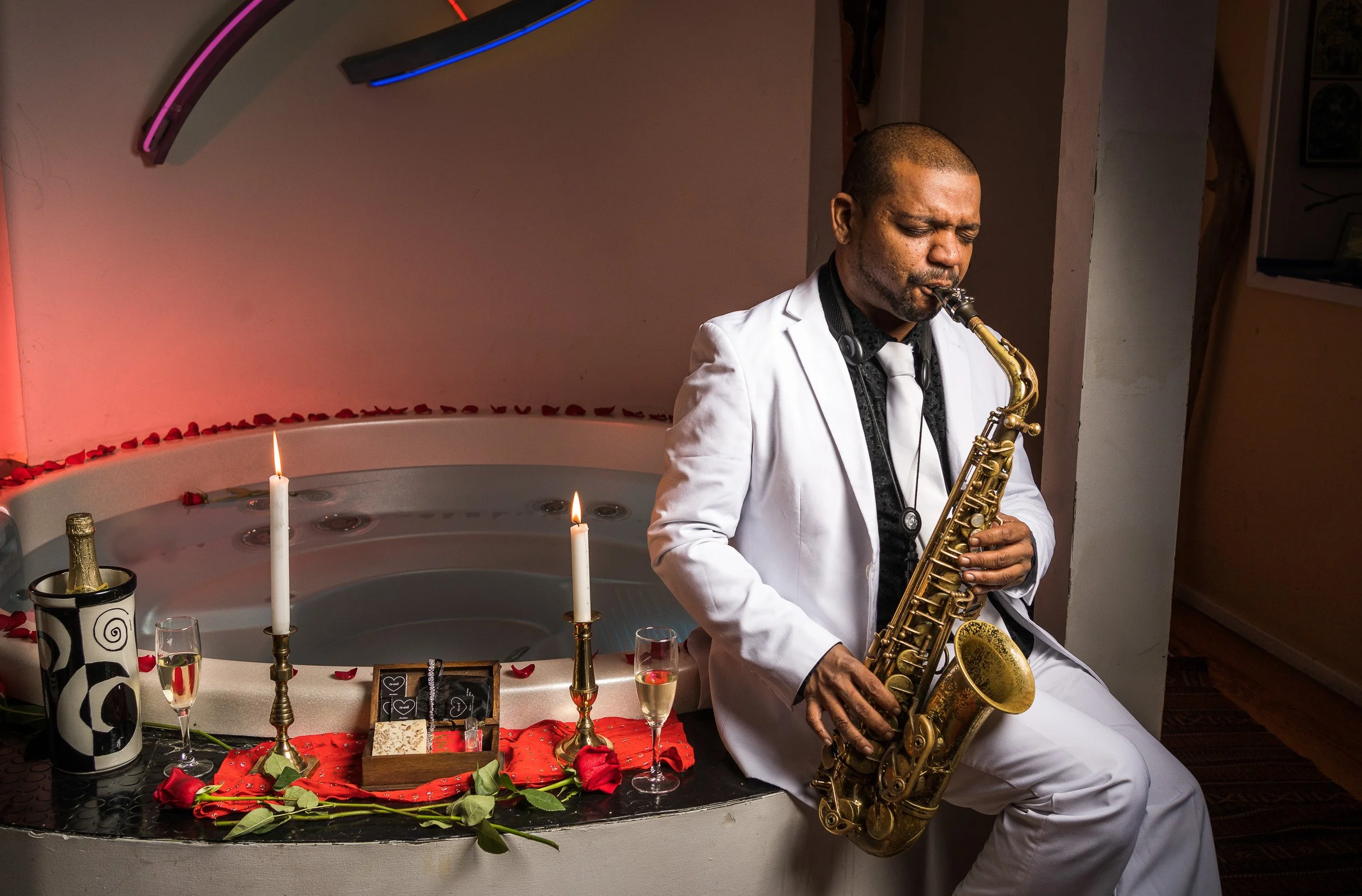 A man in a white suit playing a saxophone next to a hot tub decorated with candles, roses, and gifts, with a romantic setting.