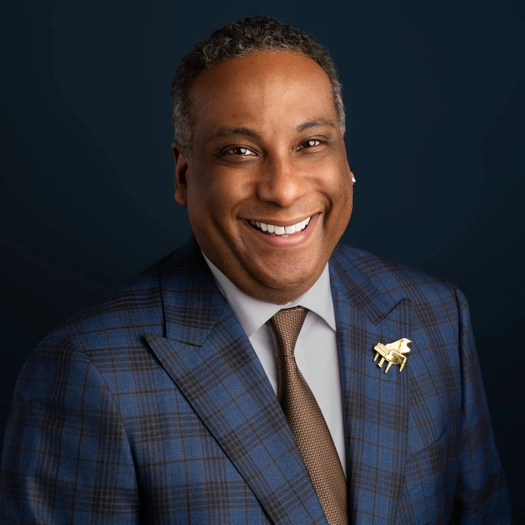 A smiling man wearing a checkered blue suit, white shirt, brown tie, and a gold ring with a design resembling a lion or similar creature.