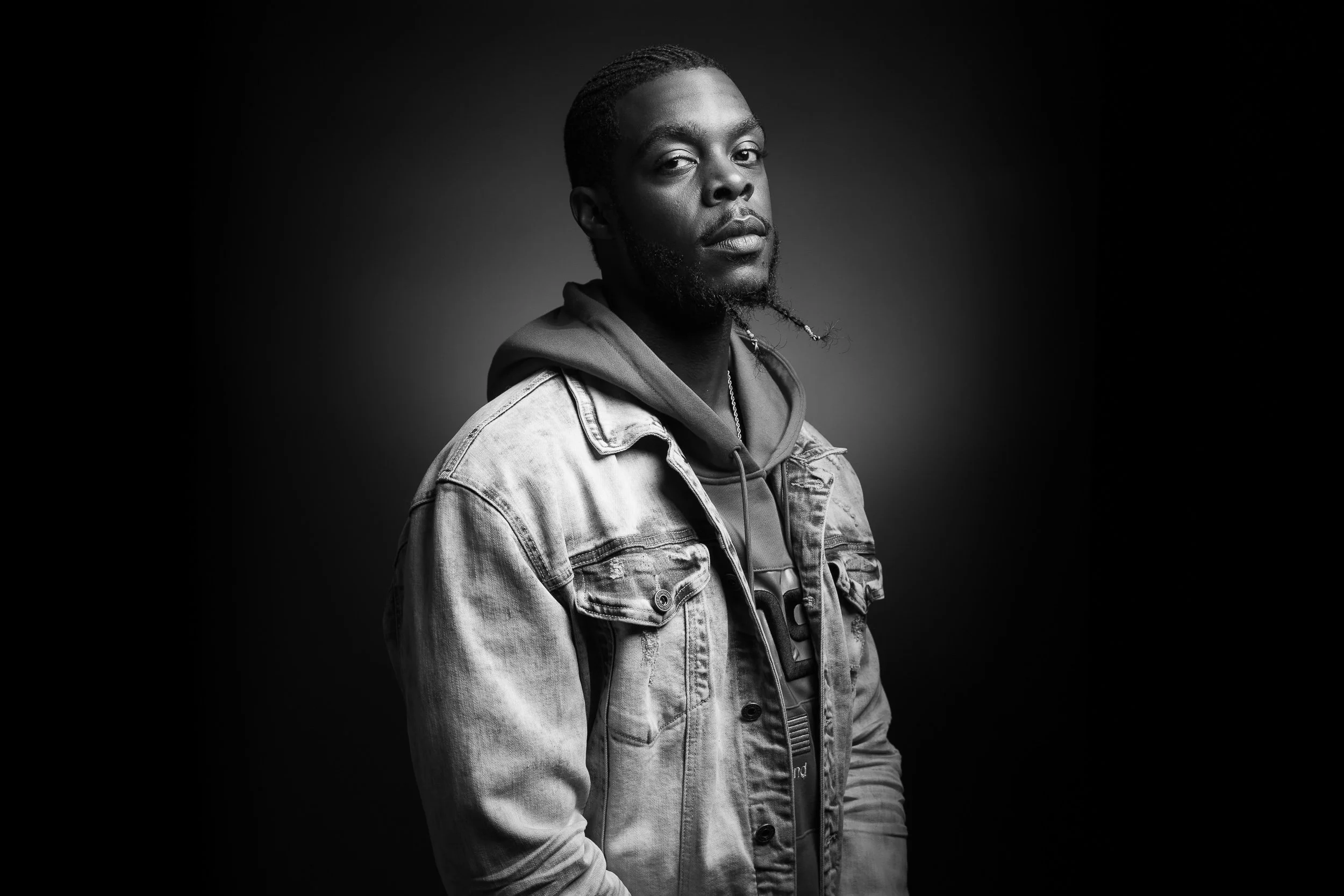 Black and white portrait of a man with short, braided hair, a beard, wearing a denim jacket and hoodie, looking at the camera against a dark background.