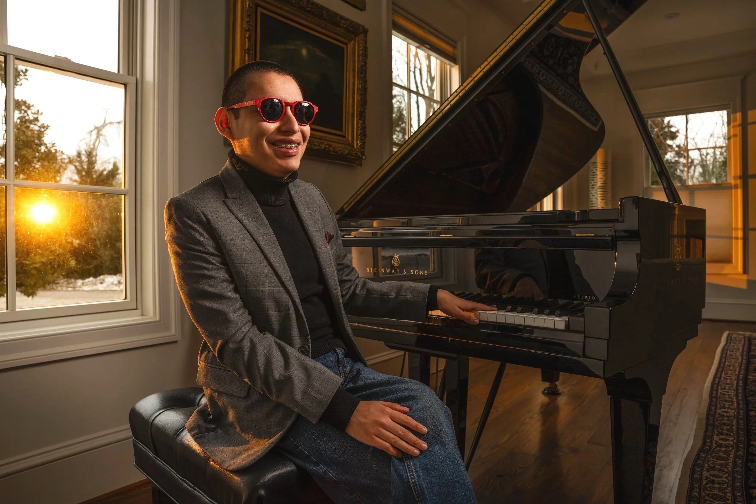 A person wearing red sunglasses and a blazer sitting by a Stefanow & Sons grand piano, smiling while the sun sets outside the window.