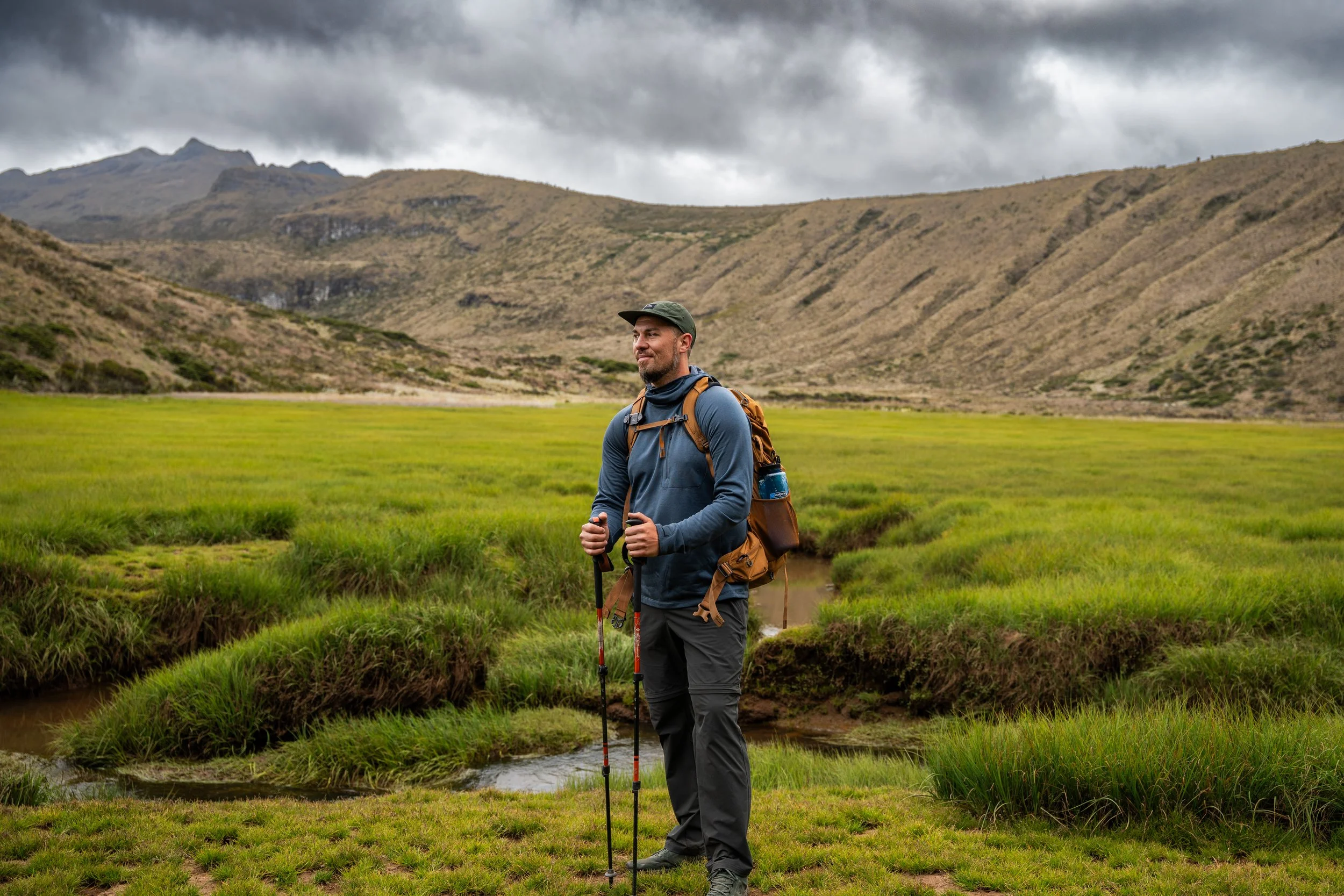 A man in outdoor hiking gear, including a gray jacket, gray pants, a hat, and a backpack, standing on a grassy area near a small stream with mountainous terrain and overcast sky in the background.