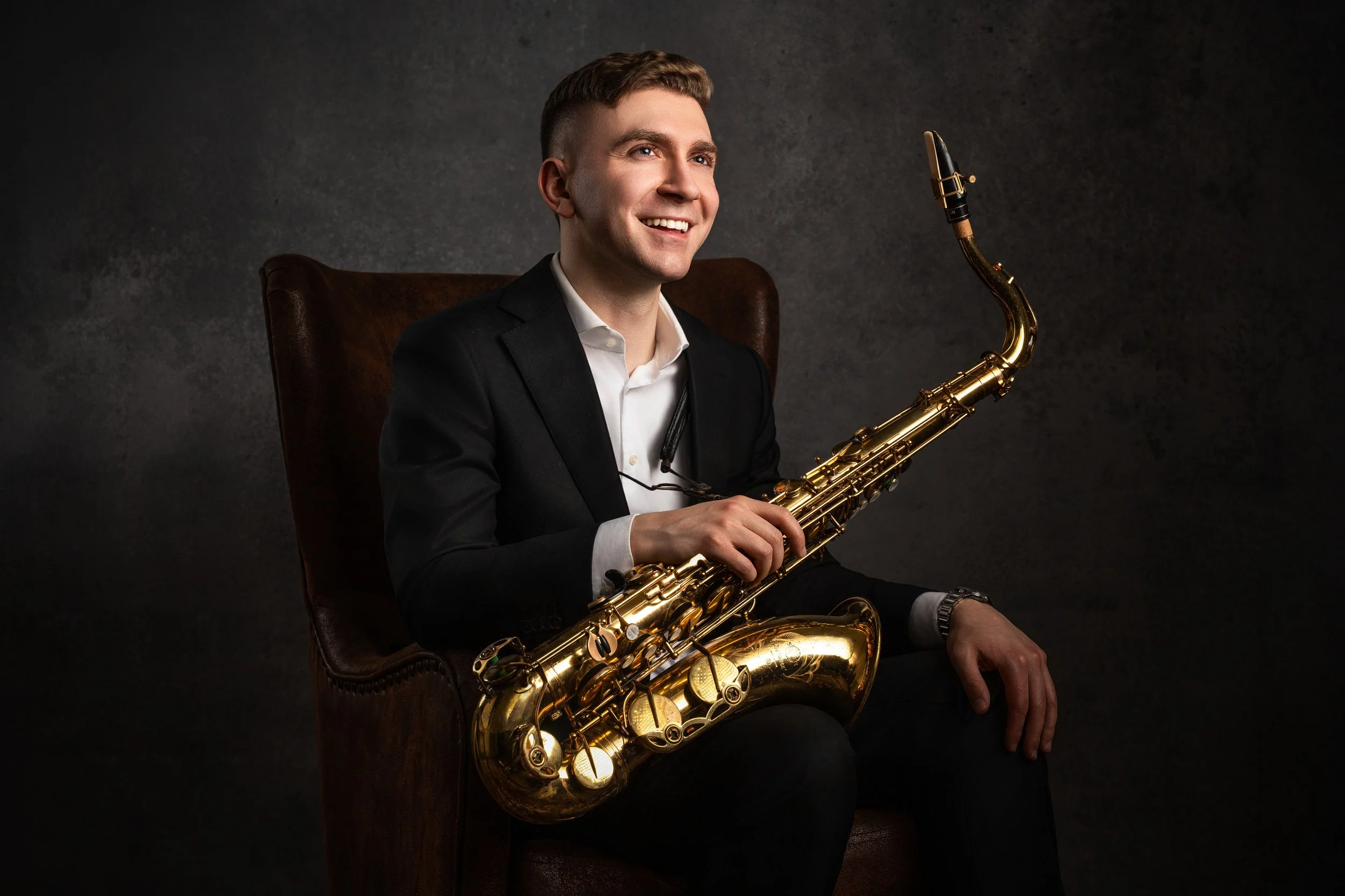 A young man dressed in a black suit with a white shirt is sitting in a brown leather chair, holding a gold saxophone, with a smile on his face, against a dark textured background.