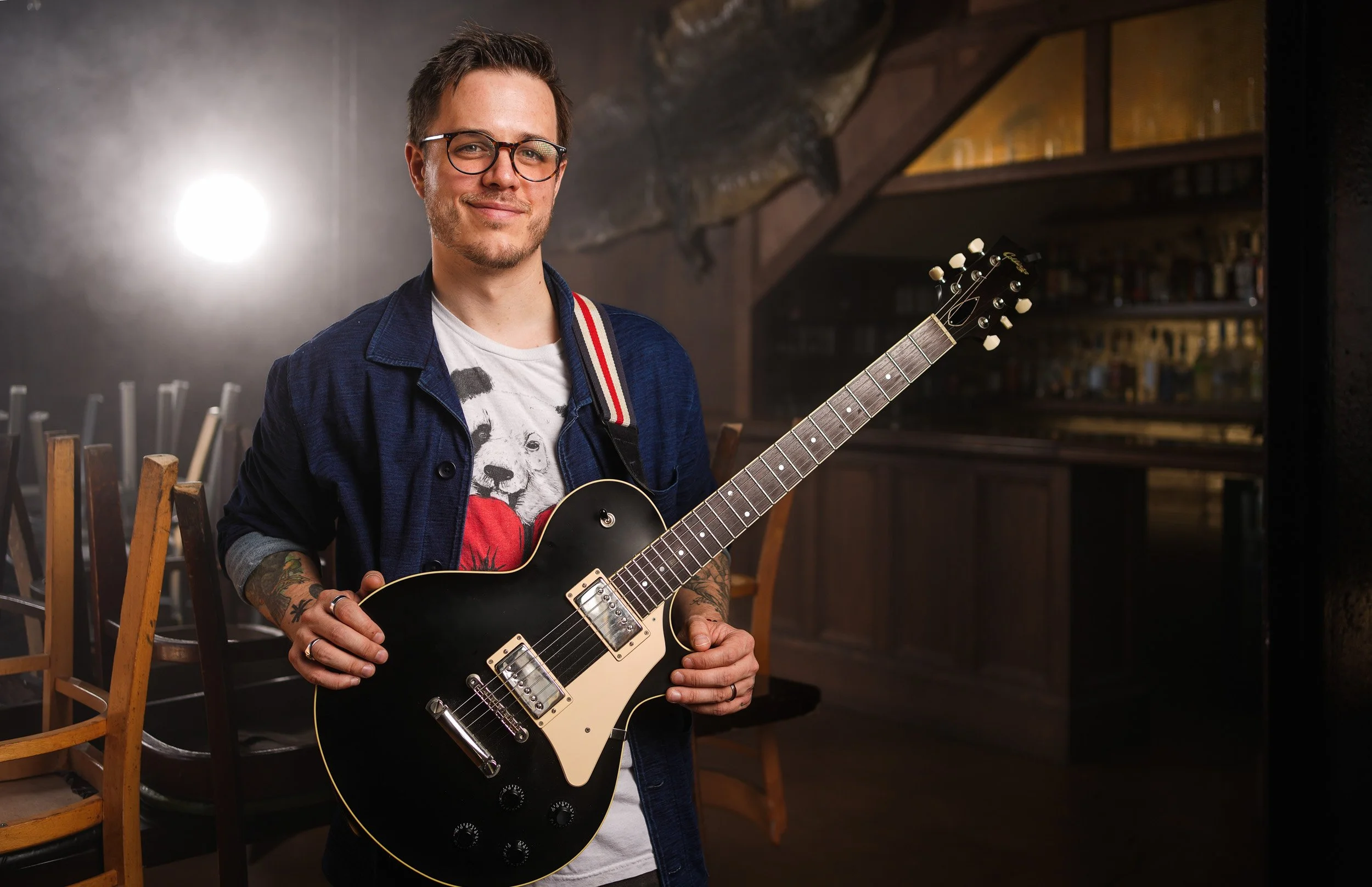A man with glasses and tattoos holding a black electric guitar in a dimly lit room with wooden chairs and shelves in the background.