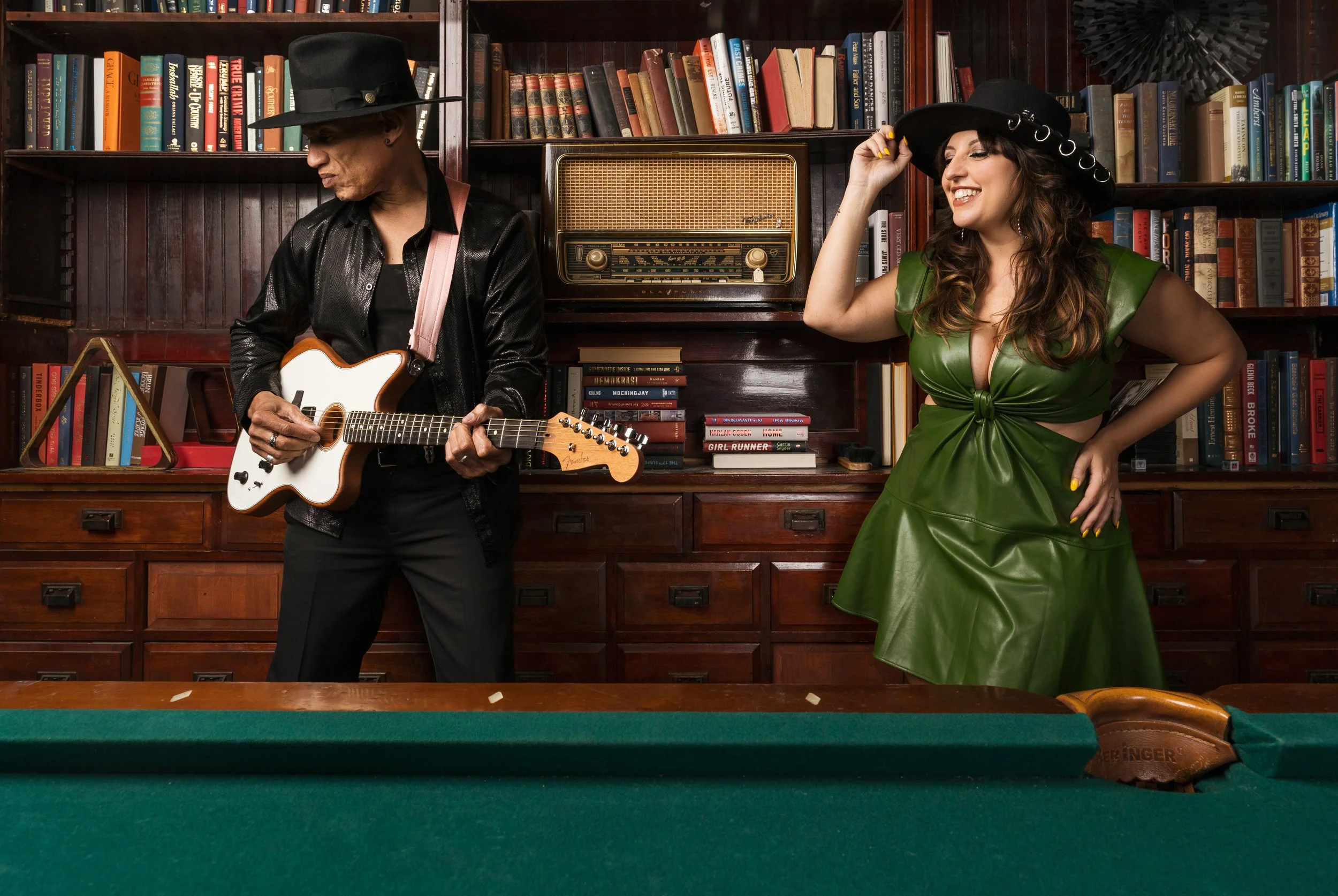 Two people, one man playing a guitar and one woman smiling and adjusting her hat, standing in front of bookshelves in a room with a vintage radio on the shelf behind them. They are both wearing hats, with the woman in a green dress and the man in a black jacket.