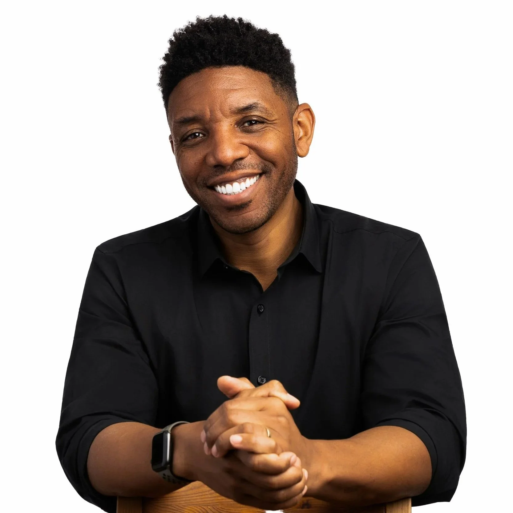 Portrait of a smiling Black man with short curly hair, wearing a black shirt and a smartwatch, sitting at a wooden table with his hands clasped.