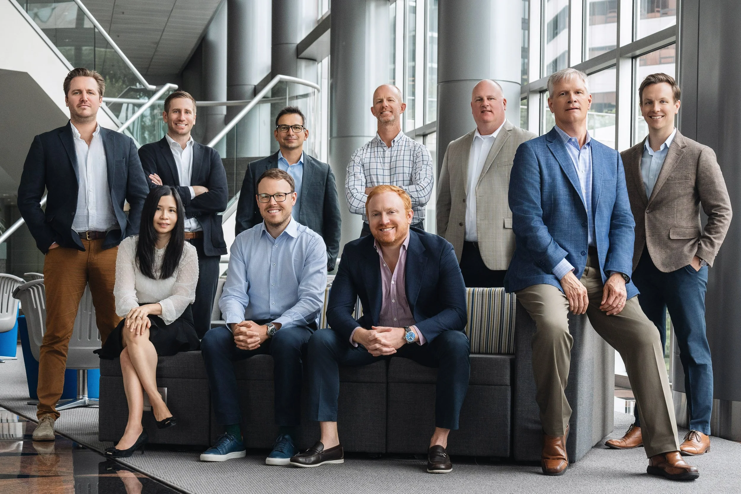 A diverse group of ten professionals, including men and women, poses together inside a modern building with large glass windows and a staircase in the background. They are dressed in business casual and formal attire, smiling and looking at the camera.