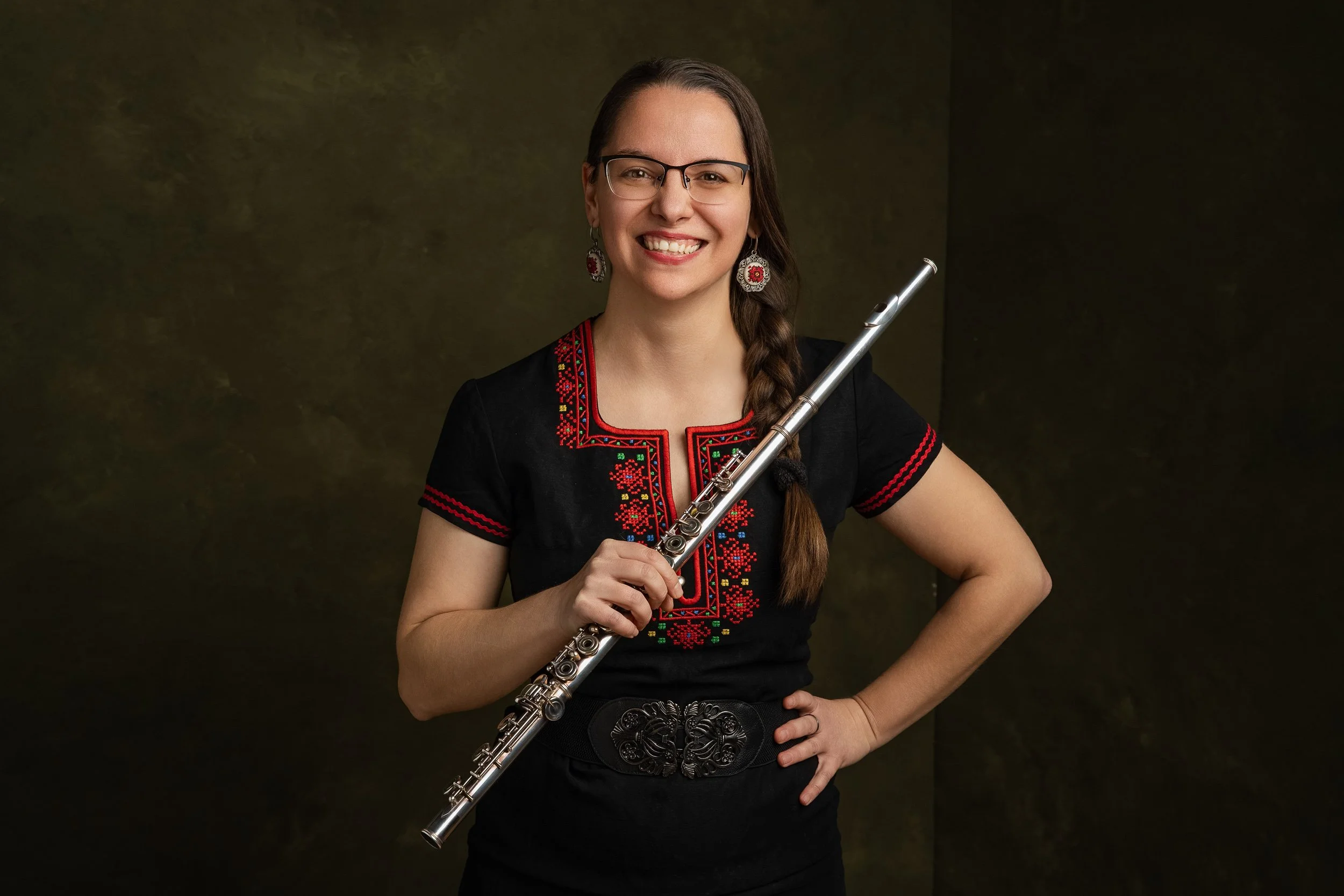 A woman wearing glasses, earrings, and a black embroidered top, smiling while holding a silver flute.
