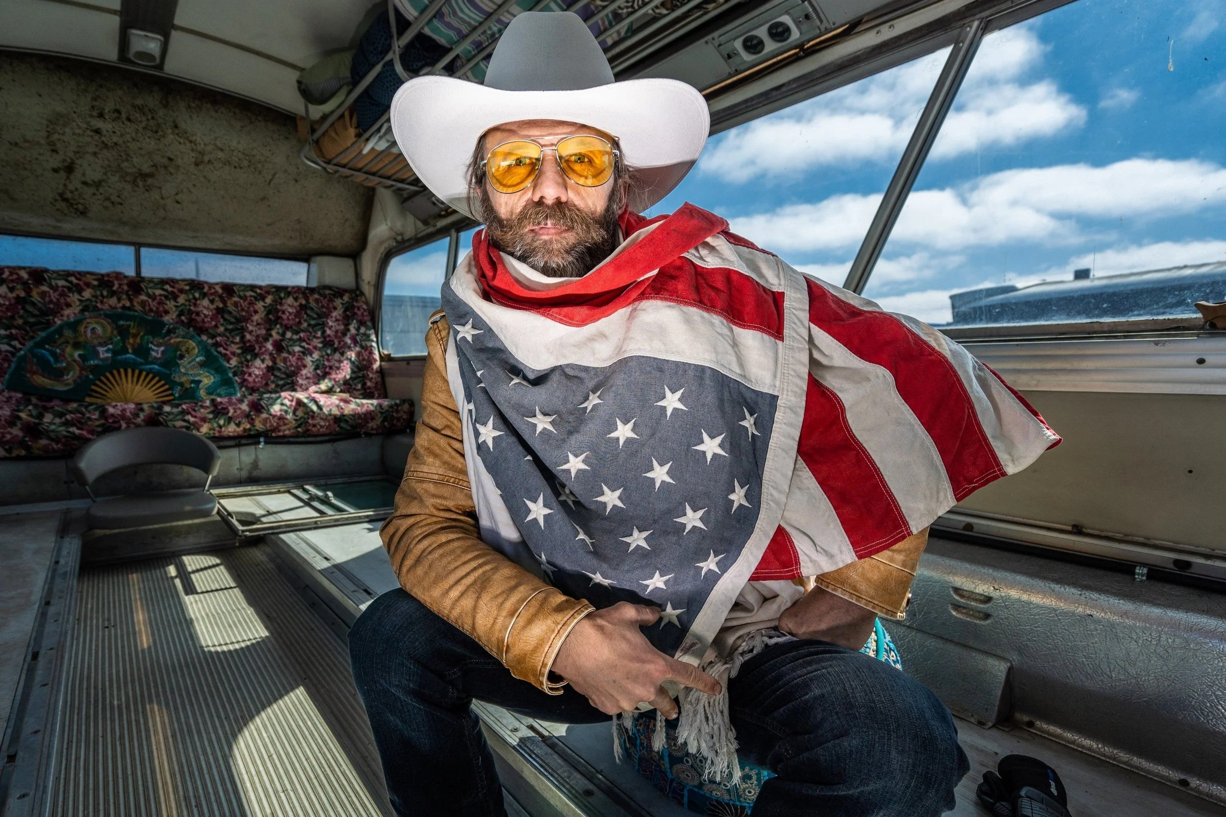 A man with a beard and glasses wearing a large white cowboy hat and an American flag bandana around his neck, sitting inside an aircraft with large windows showing a cloudy sky outside.