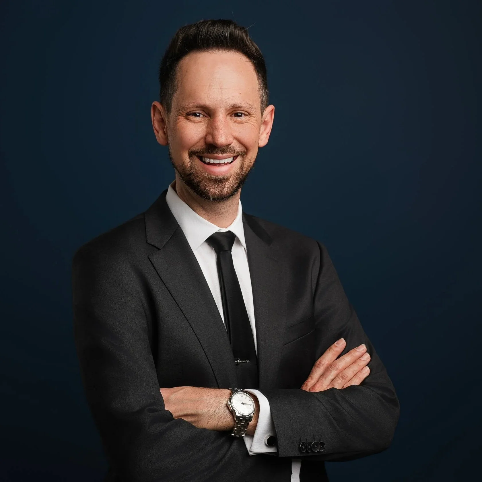 Portrait of a smiling man in a black suit, white dress shirt, black tie, wearing a wristwatch, with arms crossed, against a dark background.