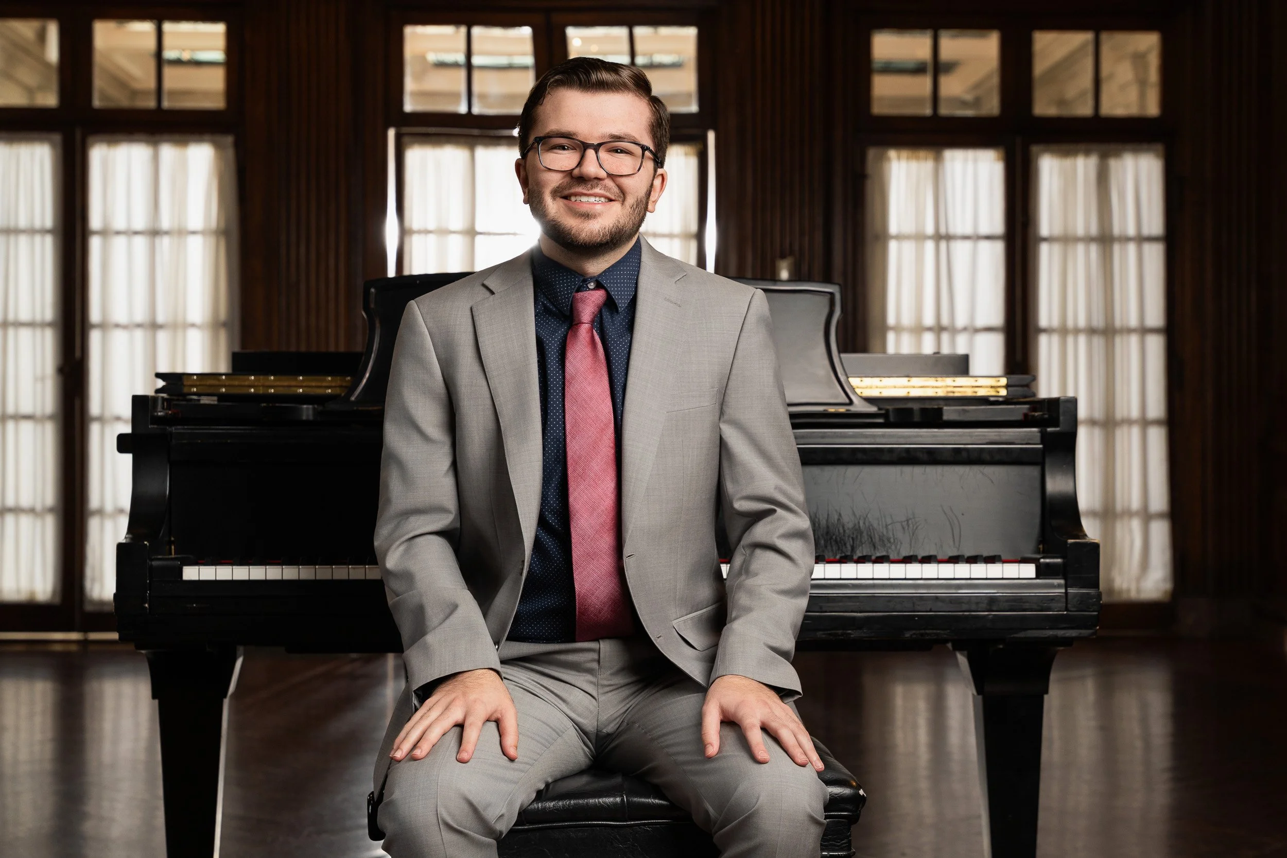 A man in a gray suit, glasses, and a red tie sitting on the edge of a black piano, smiling, in a room with tall windows and wooden paneling.