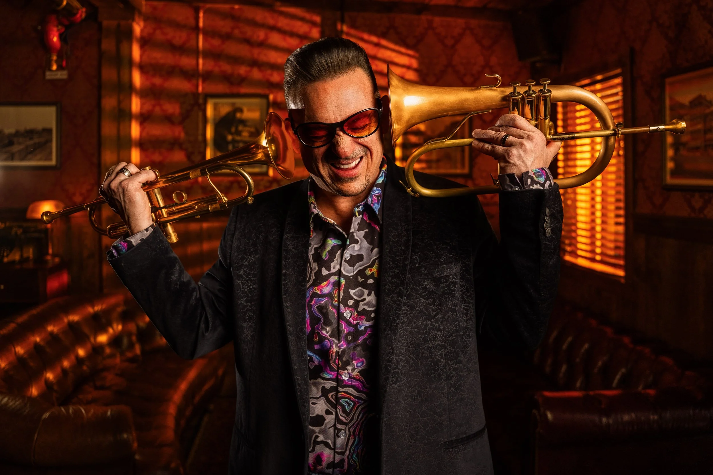 A man with styled hair, wearing sunglasses and a colorful patterned shirt, is smiling while playing a gold trumpet in a dimly lit, vintage-style room with dark red wallpaper and framed pictures on the wall.