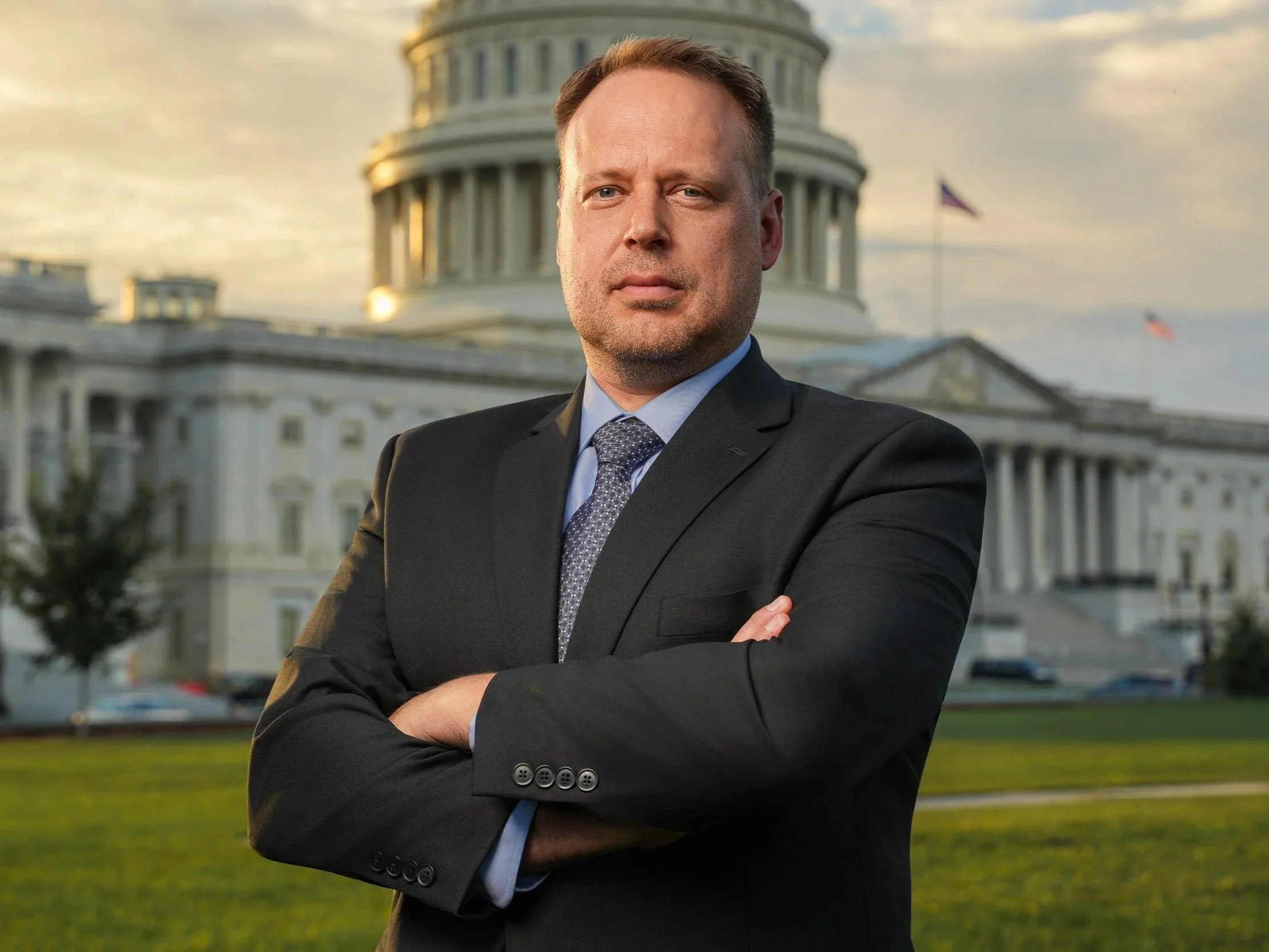 A man in a dark suit and tie standing with crossed arms in front of the U.S. Capitol building during sunset, looking confidently at the camera.