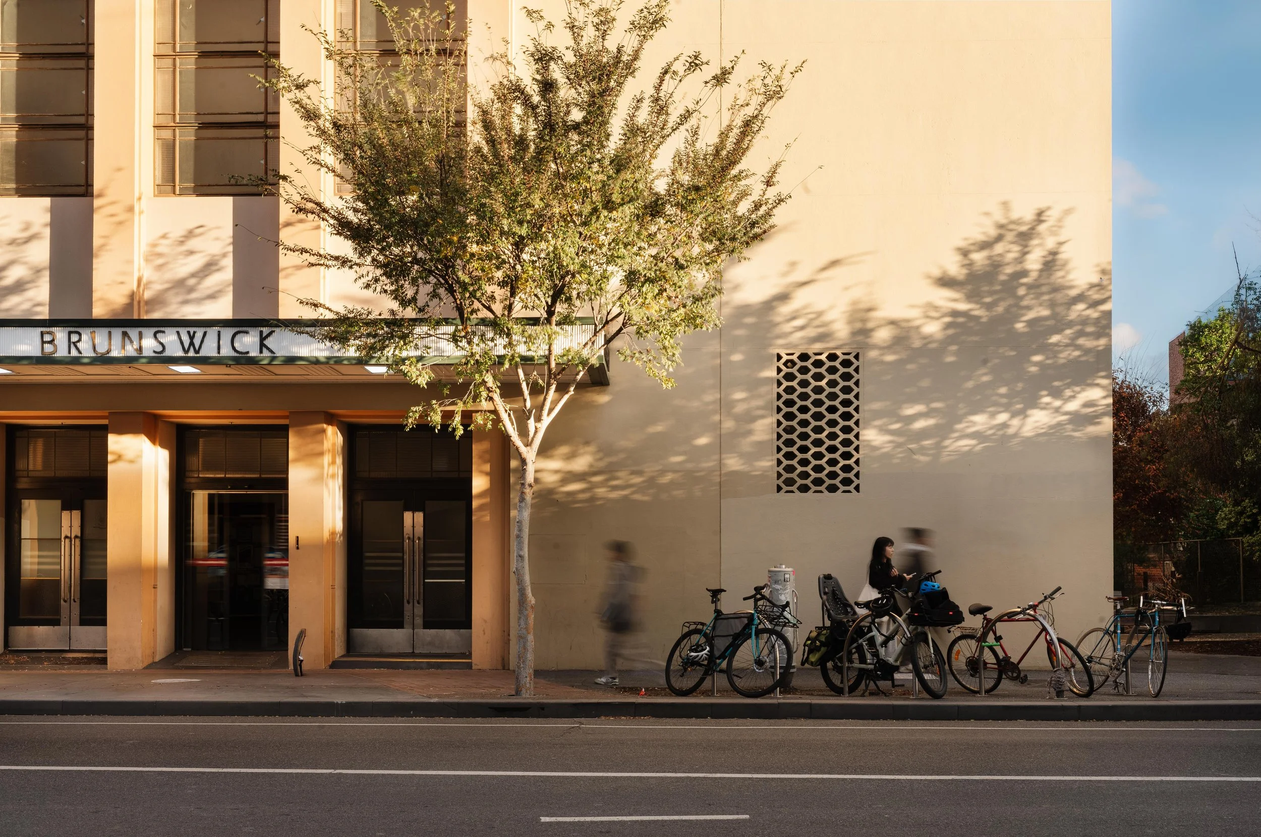 A street scene at sunset with a beige building labeled 'BRUNSWICK'. A leafy tree partially shadows the building. Bicycles are parked against the sidewalk, and a few people are walking past. The sky is clear with a few clouds.