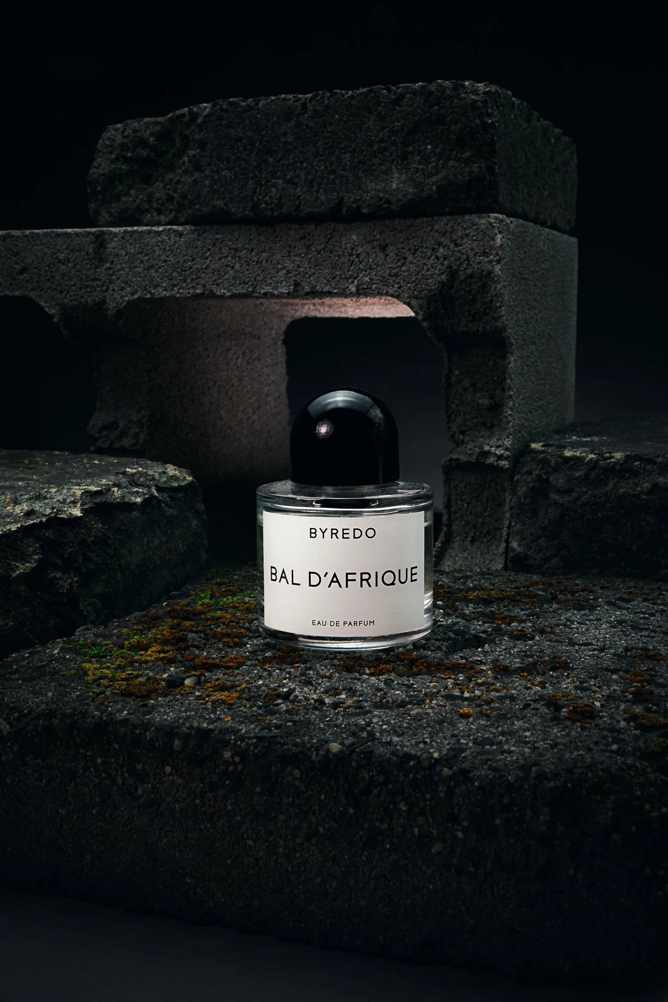 A perfume bottle labeled BYREDO BAL D'AFRIQUE in front of stacked dark volcanic rocks, with a dark background.