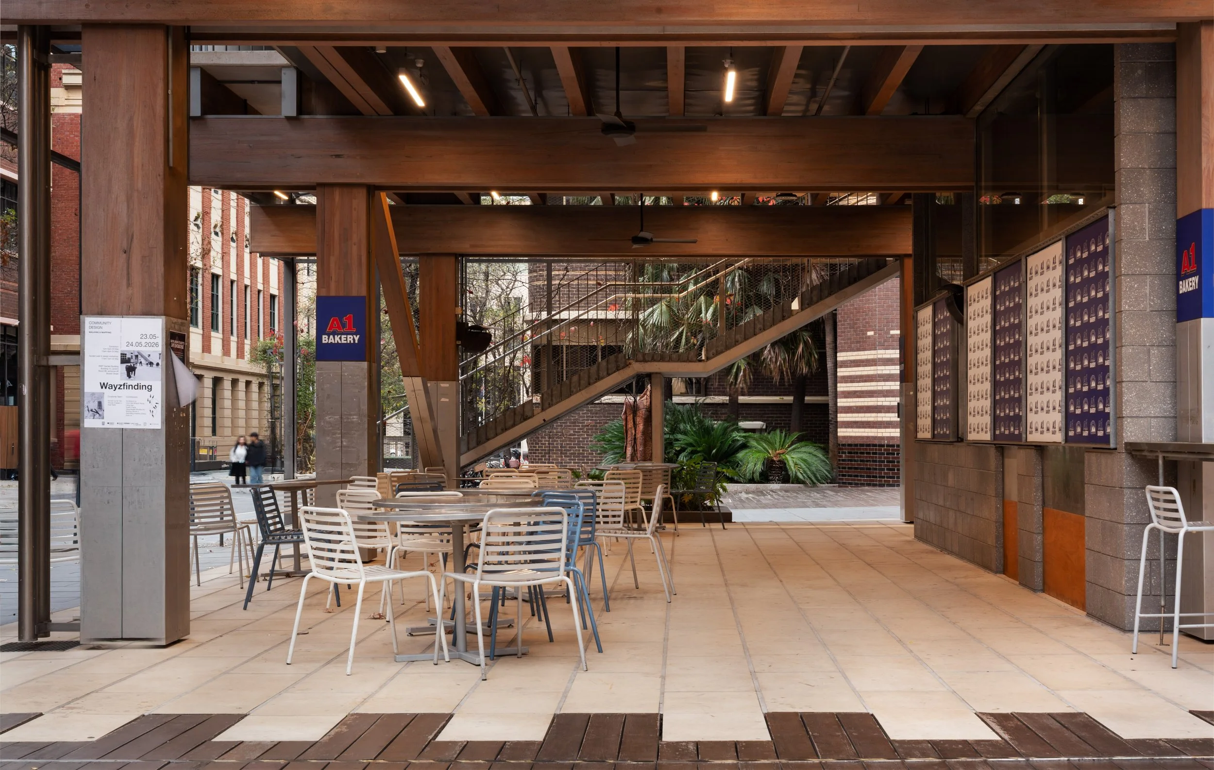 RMIT outdoor library courtyard with A1 bakery. White and black chairs and round tables, some shaded by a wooden structure with a staircase and greenery in the background.