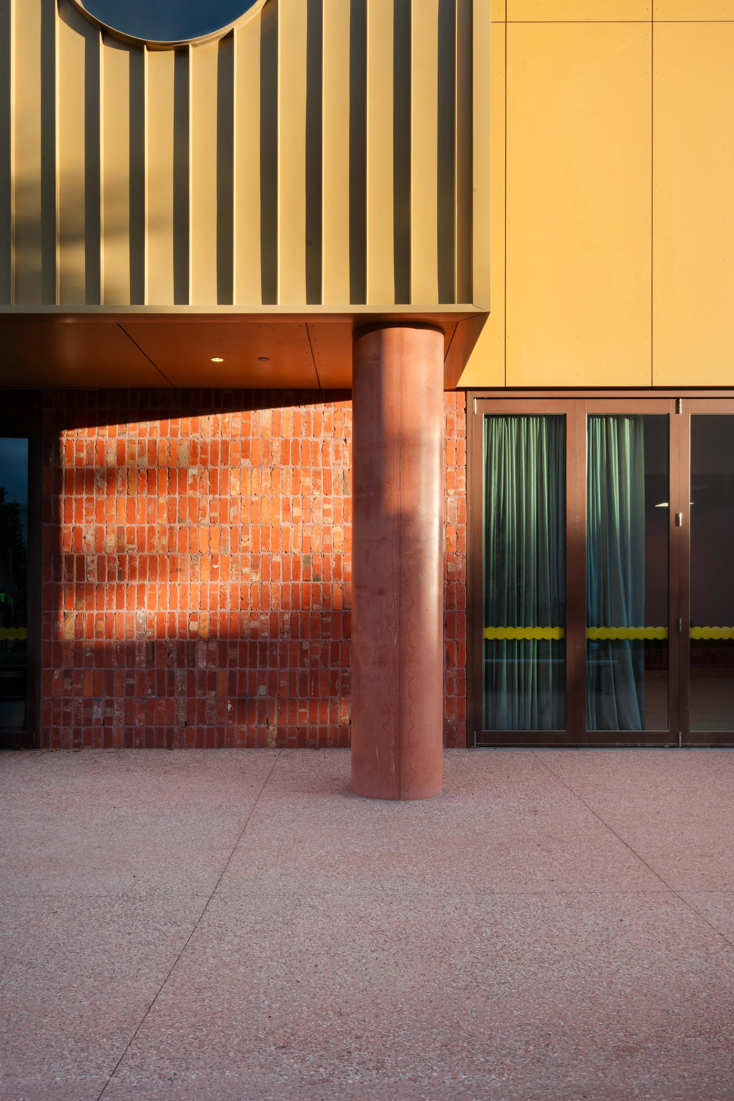 Balam Balam Place, Brunswick. Kenedy Nolan. Modern building exterior with yellow panels, brick wall, a pinkish column, and glass door with green curtains.