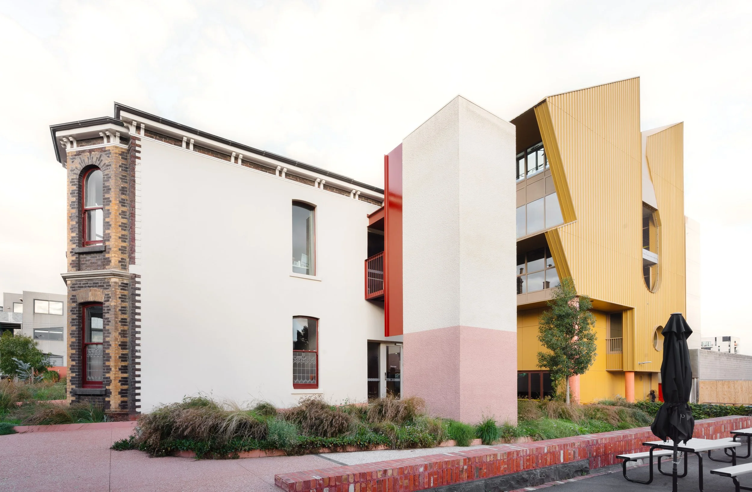 Modern building with colorful yellow, pink, white, and brick design elements and curved architectural features, with outdoor seating and greenery in the foreground.