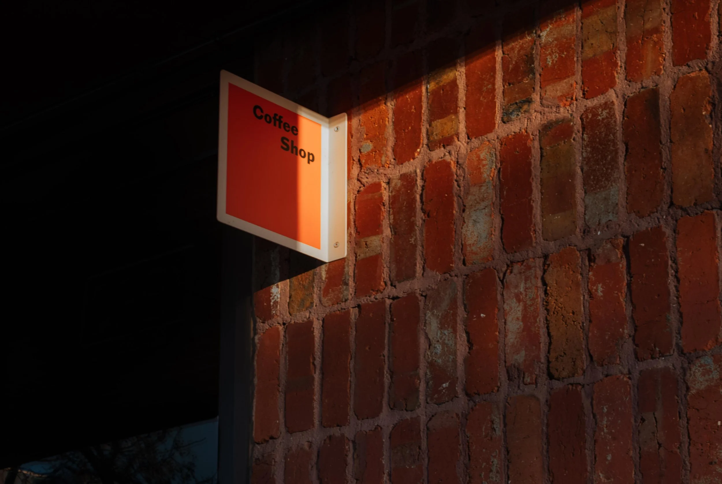 A red and white illuminated sign reading 'Coffee Shop' mounted on a brick wall, with sunlight partially casting a shadow across the sign.