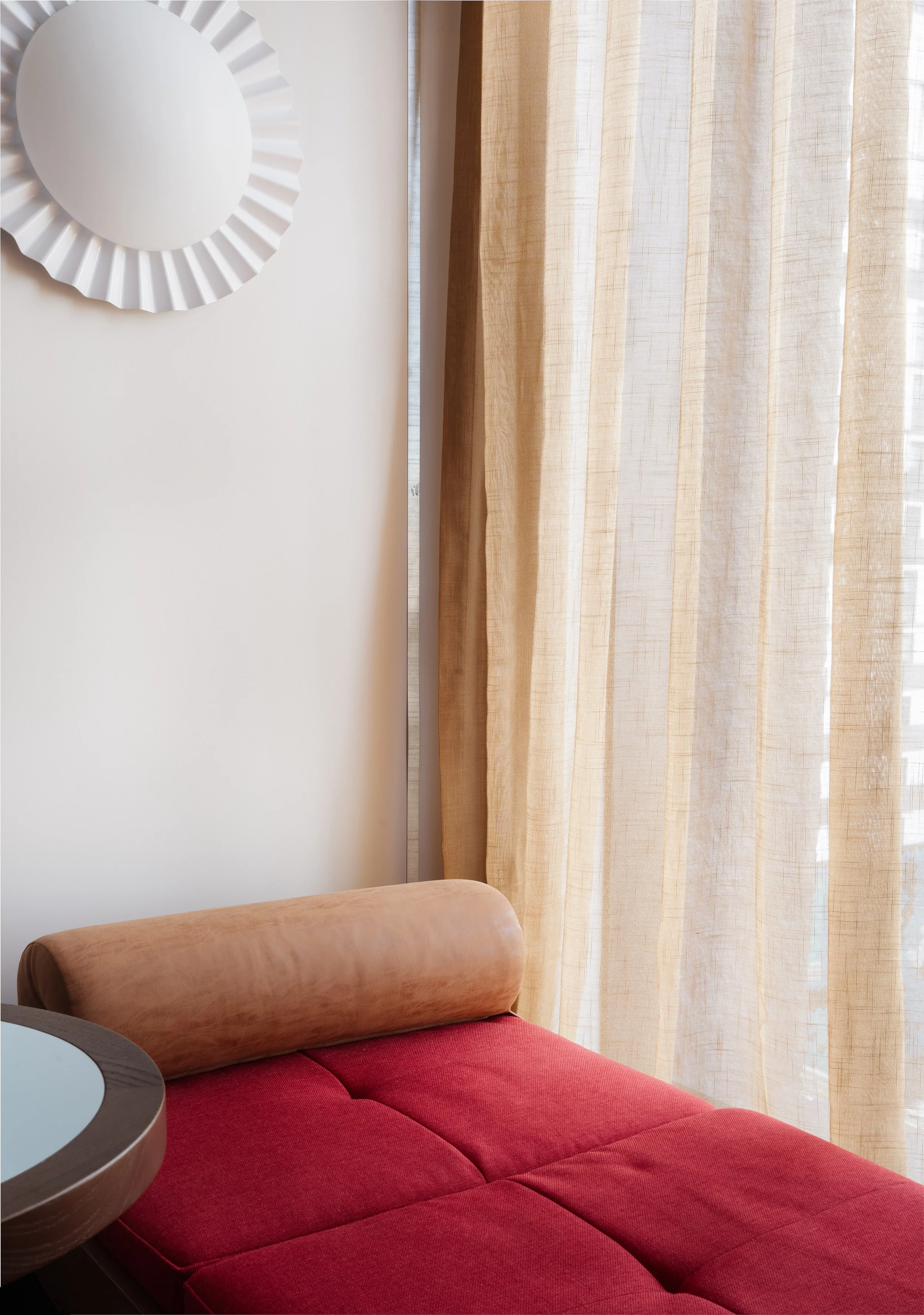 Hannah St Hotel Photography. A corner of a hotel room with a red cushioned sofa, a wooden armrest, beige curtains, and part of a white wall with a decorative white circular mirror.