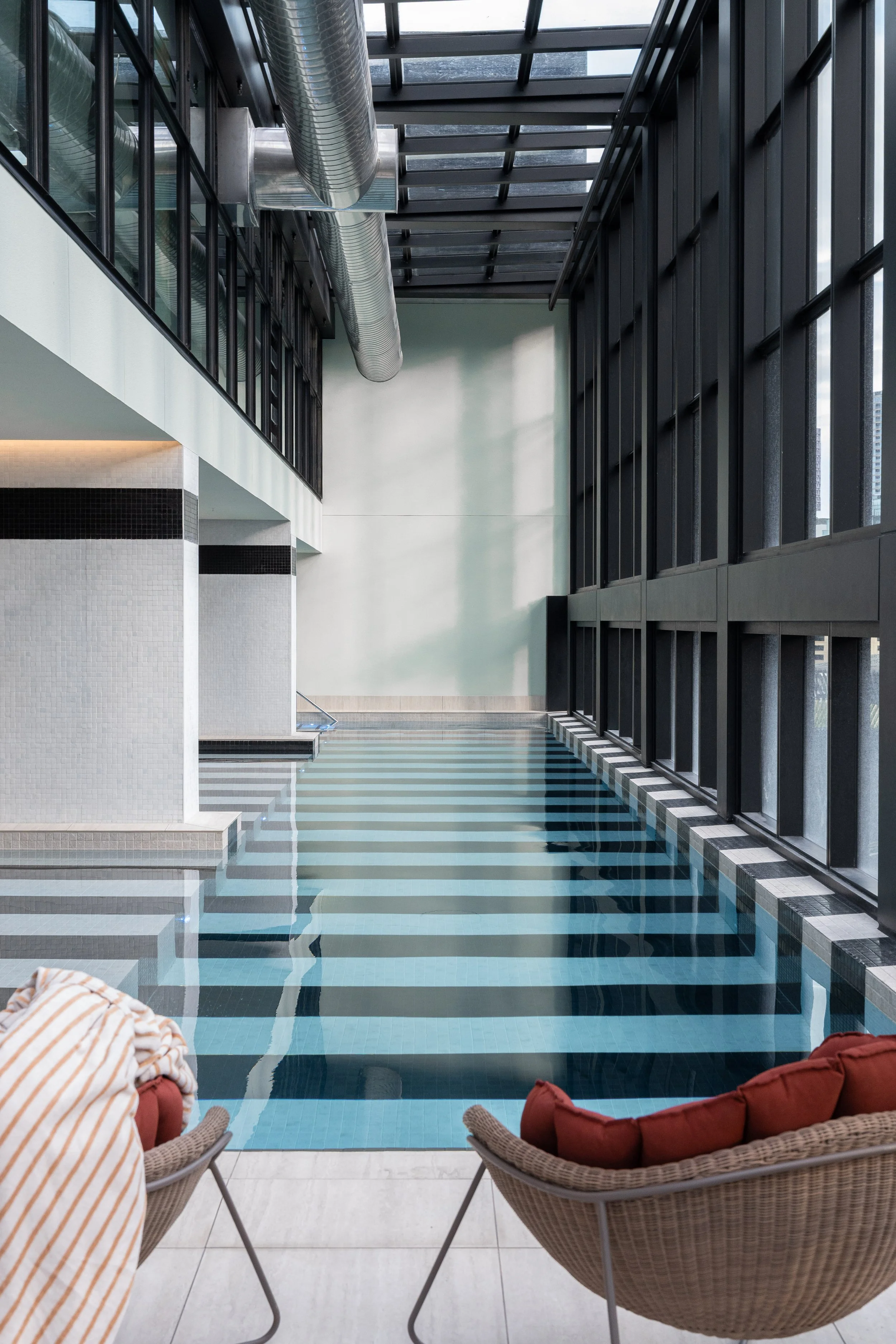 Indoor swimming pool area with black and light blue tiled floor, wicker chairs with cushions, large windows, and exposed ductwork on the ceiling.