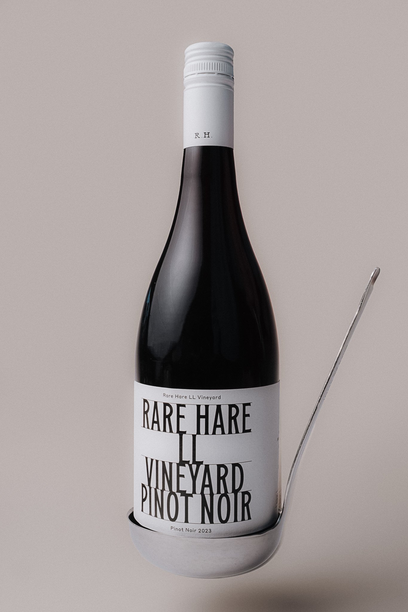 Rare Hare wine bottle with a white label featuring black text, reading 'RARE HARE VINEYARD PINOT NOIR,' placed inside a white circular holder with a cutout for the neck, and a metal tool inserted beside it against a plain background.