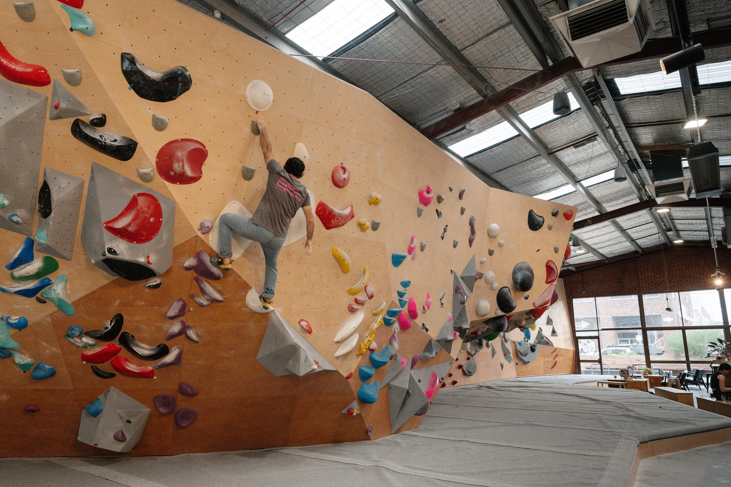 Northside Boulders Indoor bouldering gym with a person climbing on a wooden wall with colourful holds, exposed ceiling with industrial lighting, and large windows. Shot for Vertical Ascents