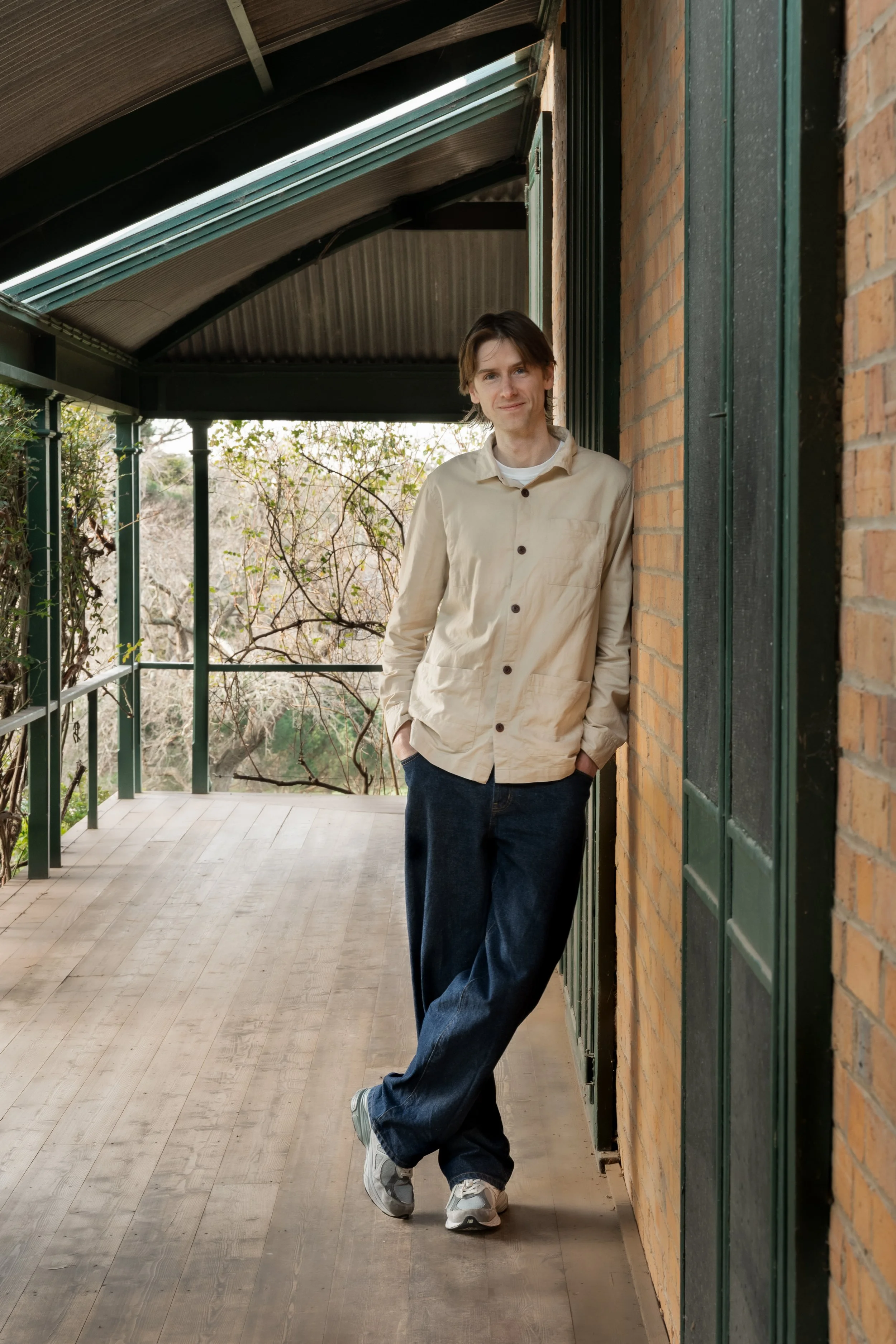 A young man with brown hair smiling and leaning against a brick wall on a covered outdoor porch, wearing a beige button-up shirt, dark jeans, and gray sneakers.