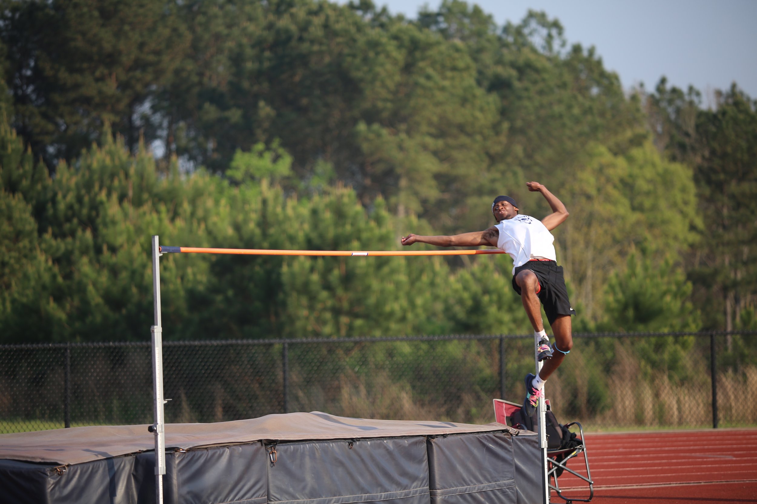 A male athlete in a white tank top and black shorts leaps over a high jump bar on an outdoor track field during daytime, with green trees in the background.