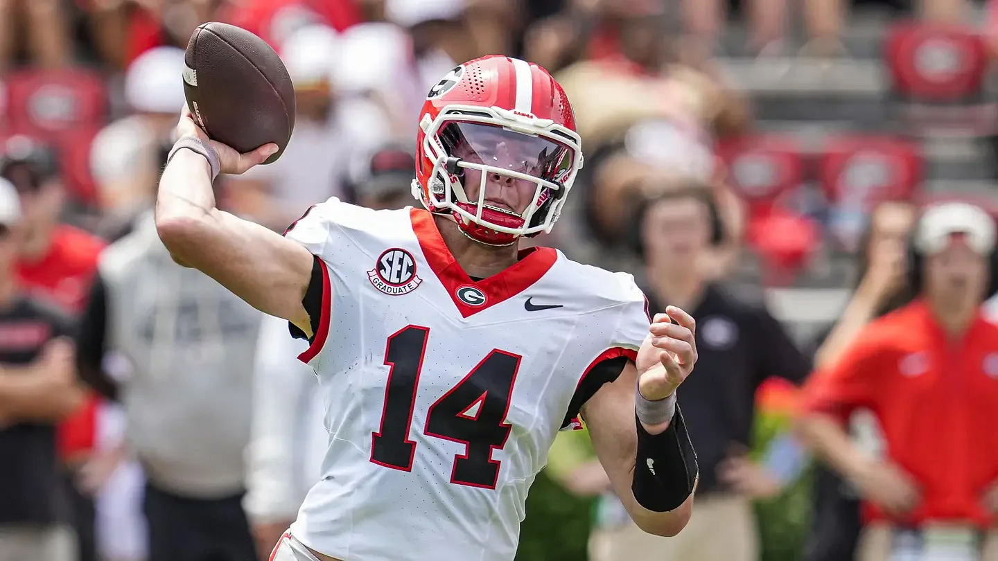 American football player wearing a red helmet and white jersey with the number 14, holding a football in his right hand, on a field with spectators in the background.
