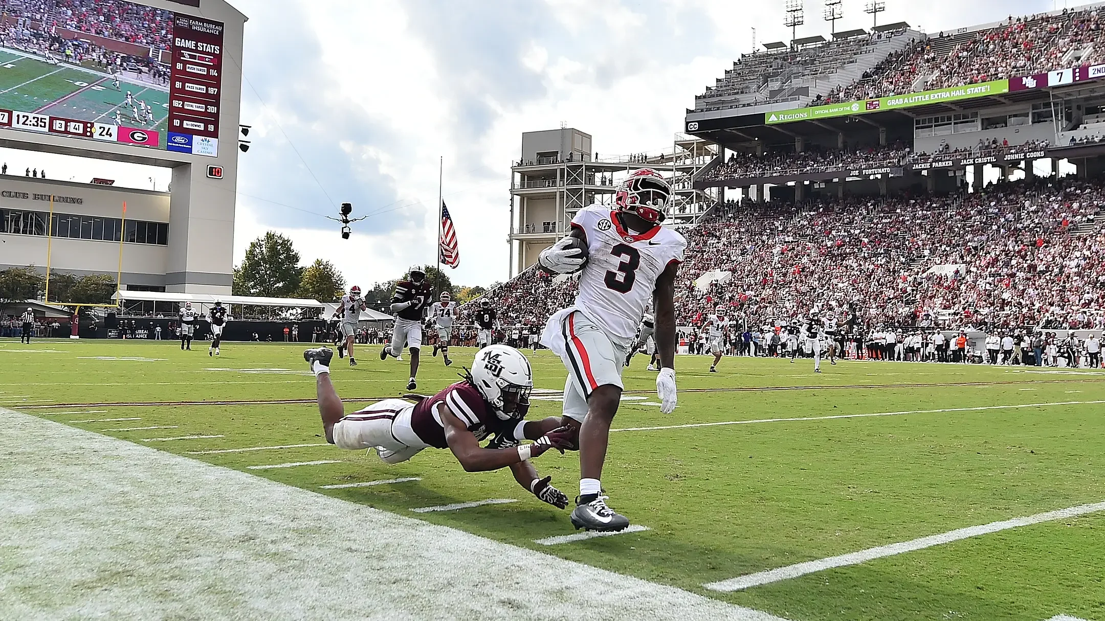 A football game in progress with a player in a white jersey and helmet running with the ball past a player in a maroon jersey who has just been tackled. The crowd is seated in the background with a scoreboard and stadium structures visible.