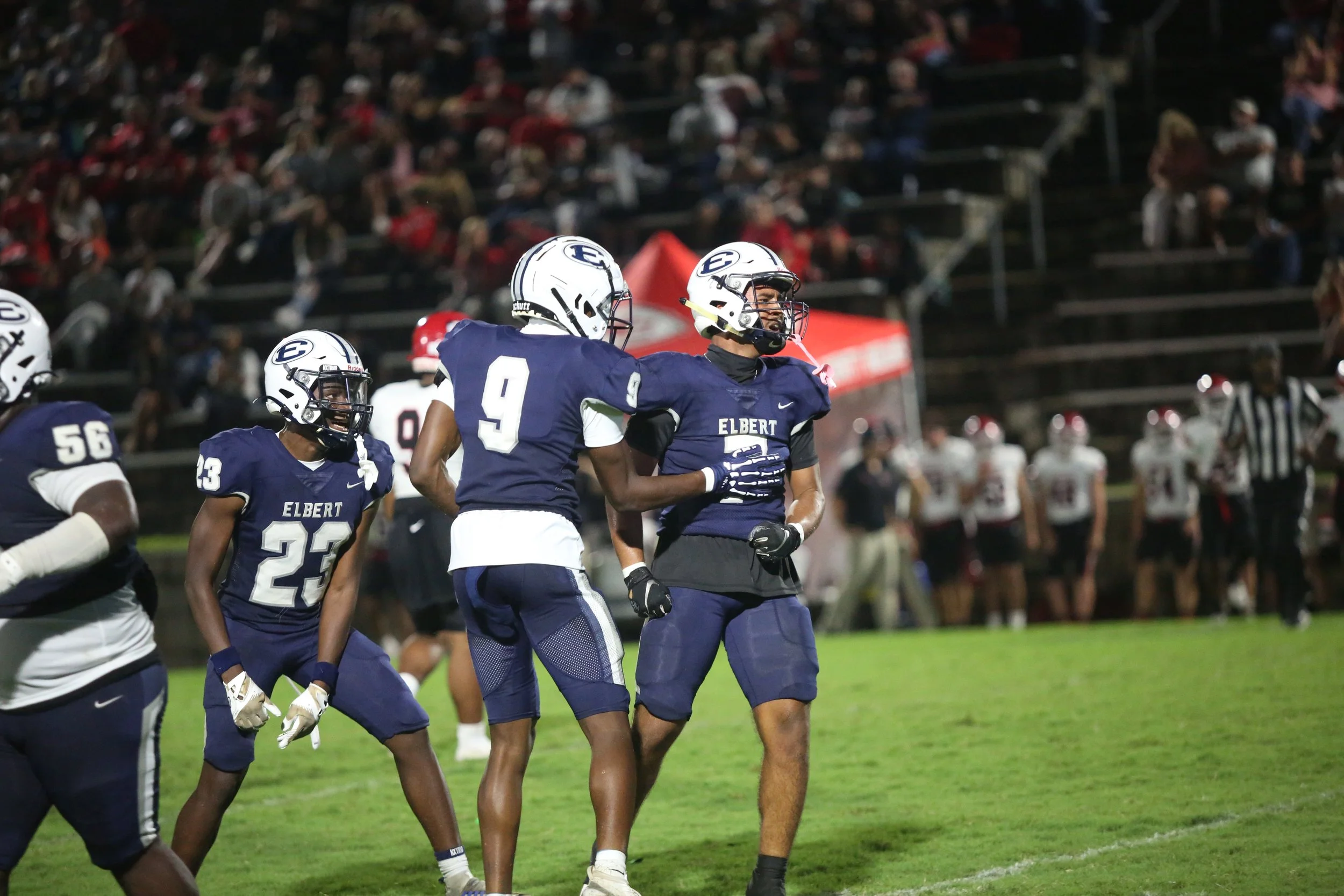 Football players wearing blue and white uniforms with 'ELBERT' on the front, on a green football field at night, celebrating and preparing for play with an audience in the background.