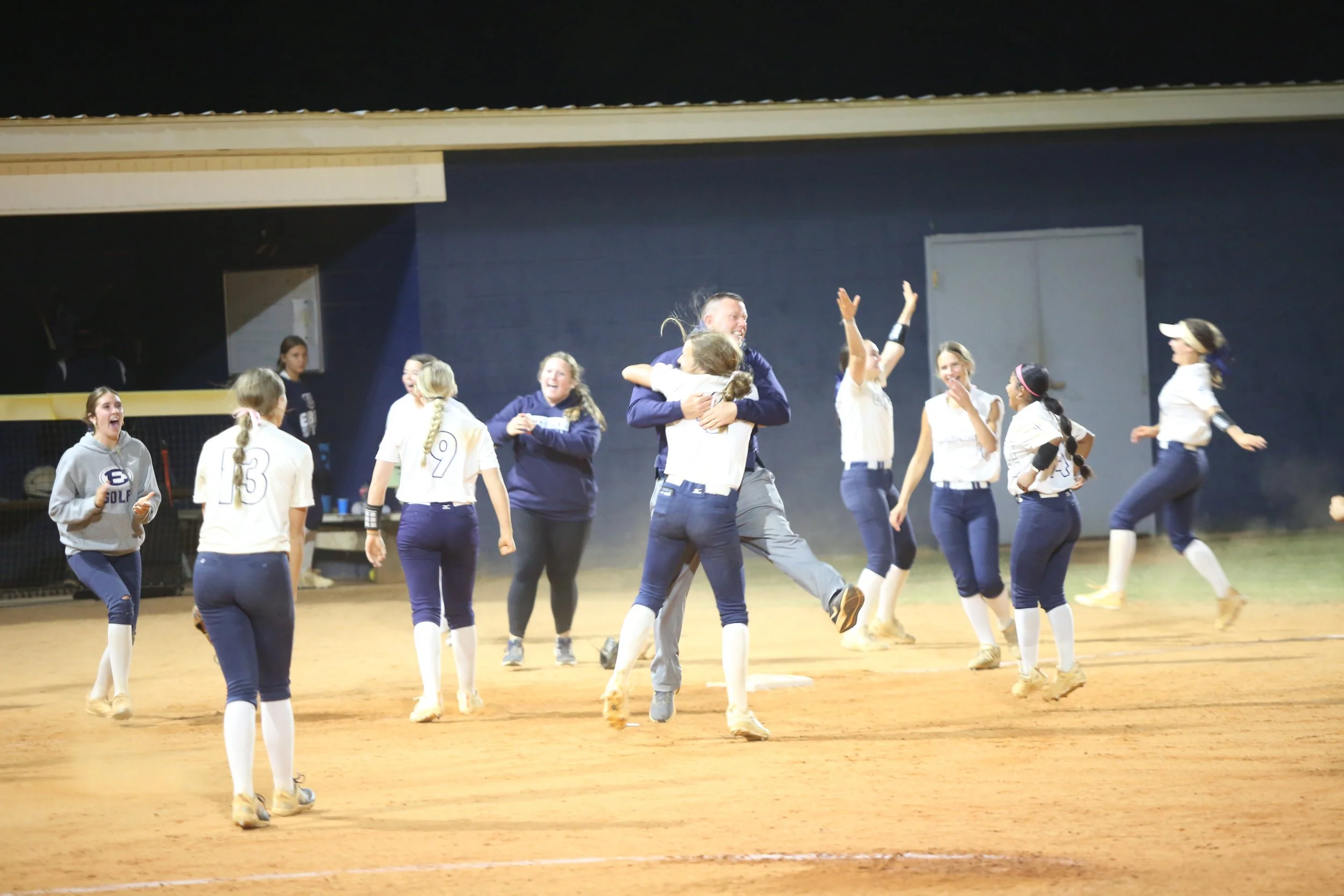 A group of young female softball players and coaches celebrating on the field at night, with some hugging and smiling.