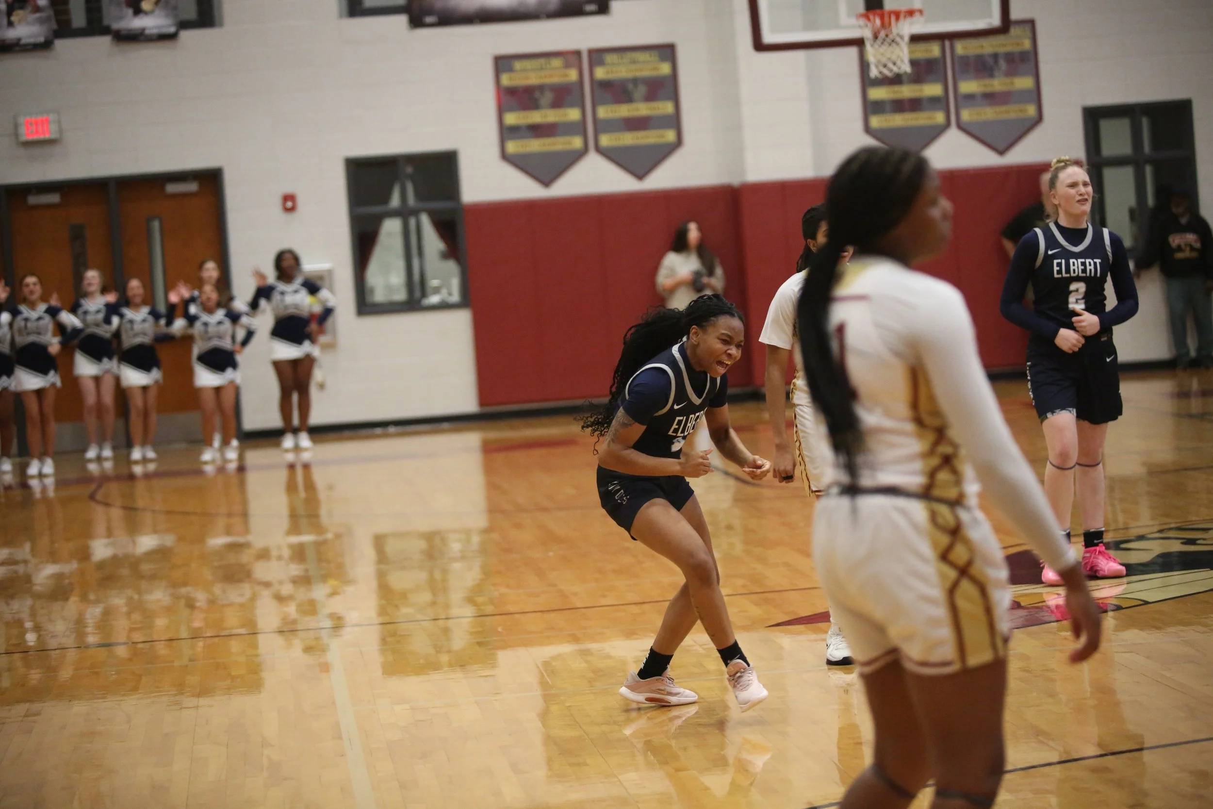 A girls' volleyball team in uniform, some with hands on hips and one with a fierce expression, playing or practicing in a gymnasium while other cheerleaders are in the background.