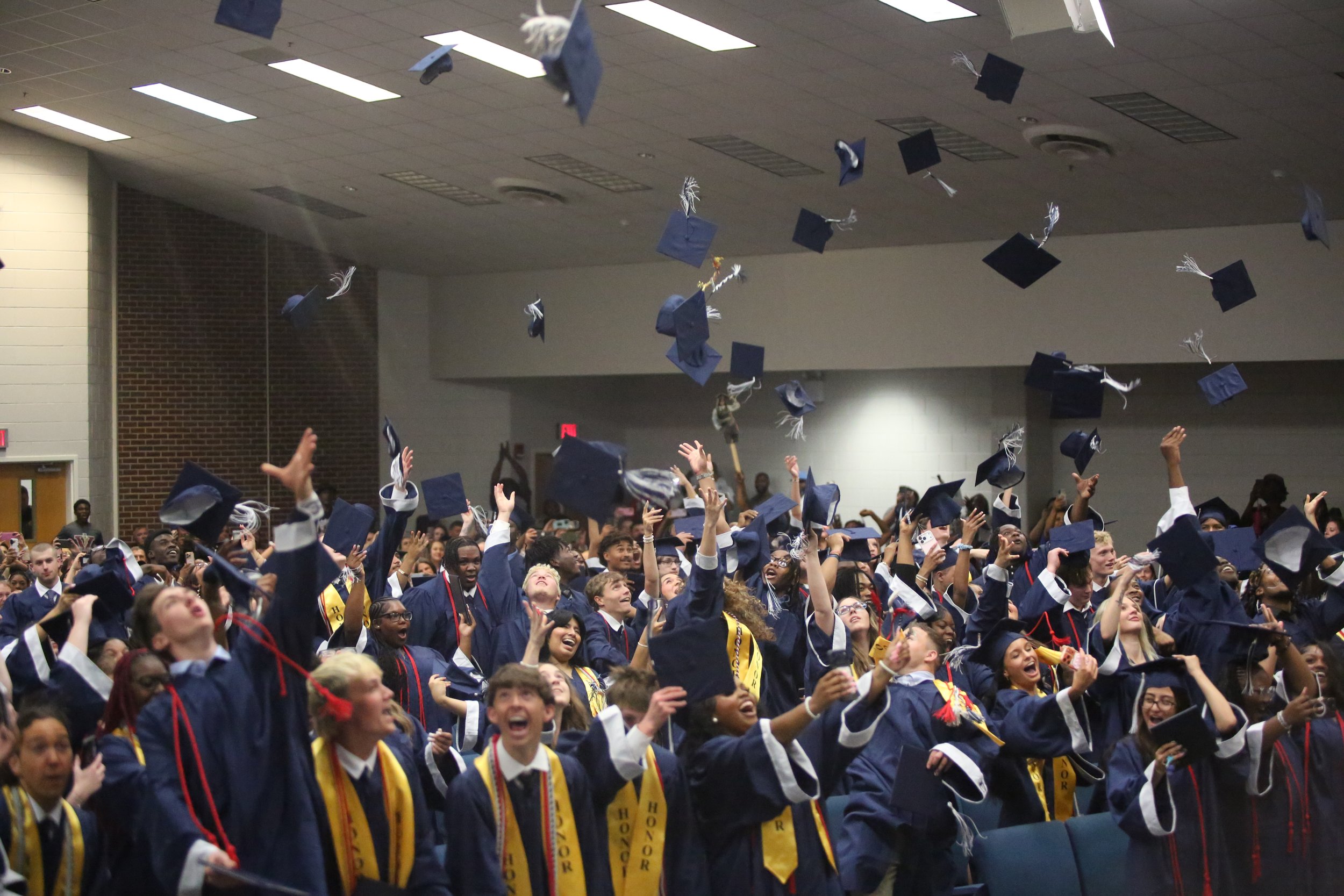 A large group of students dressed in caps and gowns celebrating at graduation, with some caps flying in the air inside a large indoor hall.