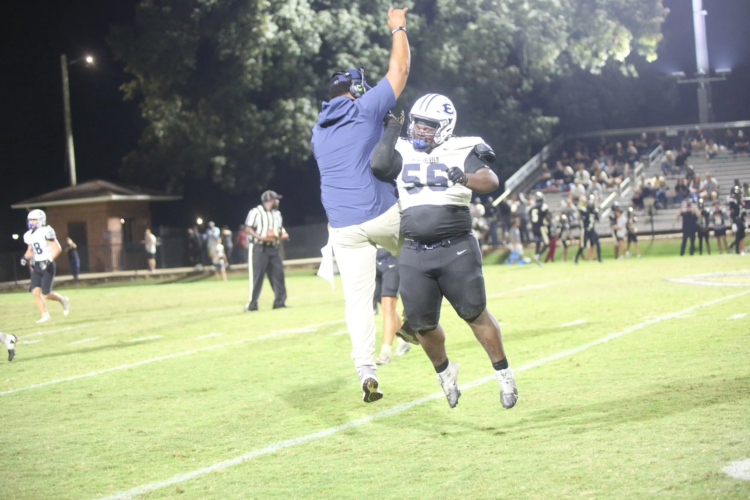 Football players celebrating on the field at night, with one player jumping and another giving a high five, audience in the stands, and trees in the background.