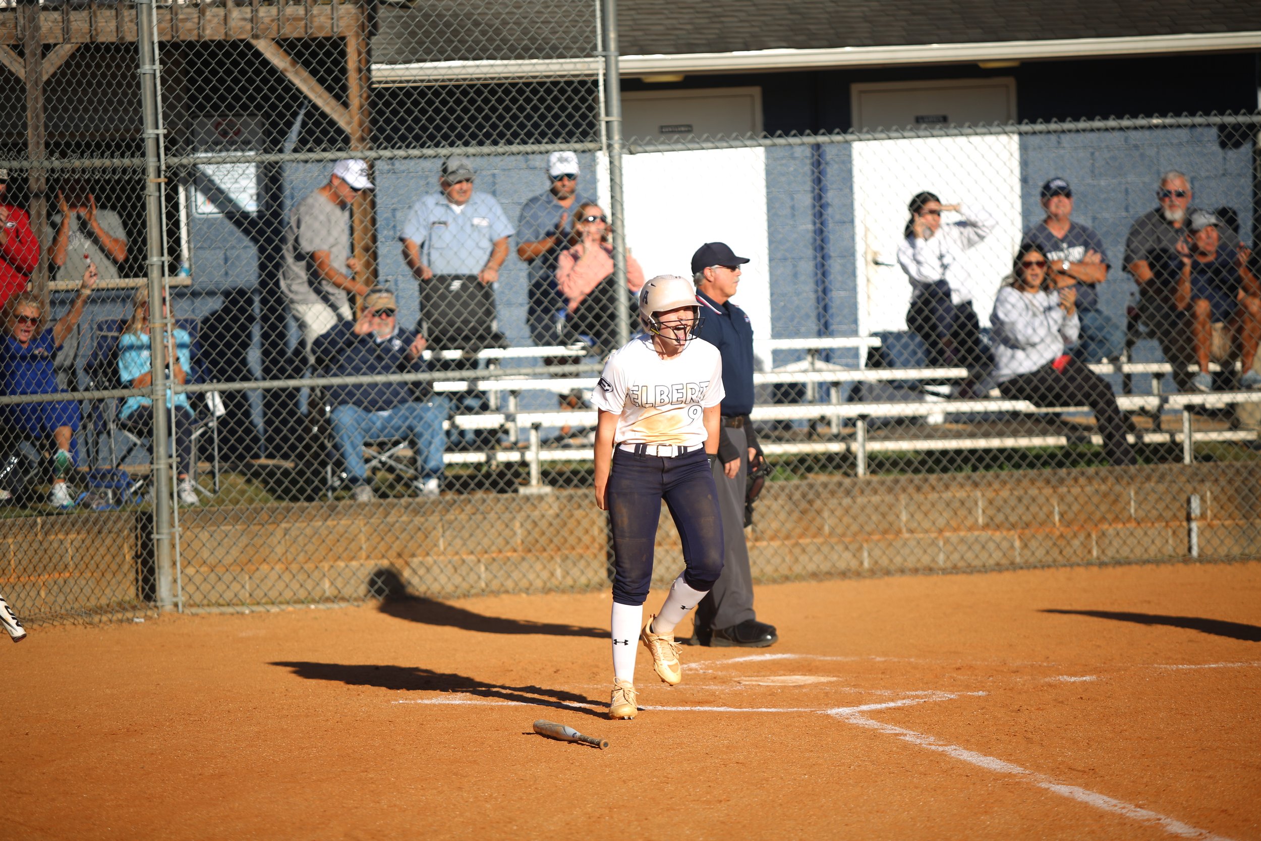 A woman in a softball uniform walking on a dirt field after hitting, with a bat on the ground nearby, while spectators watch and cheer from behind a chain link fence.