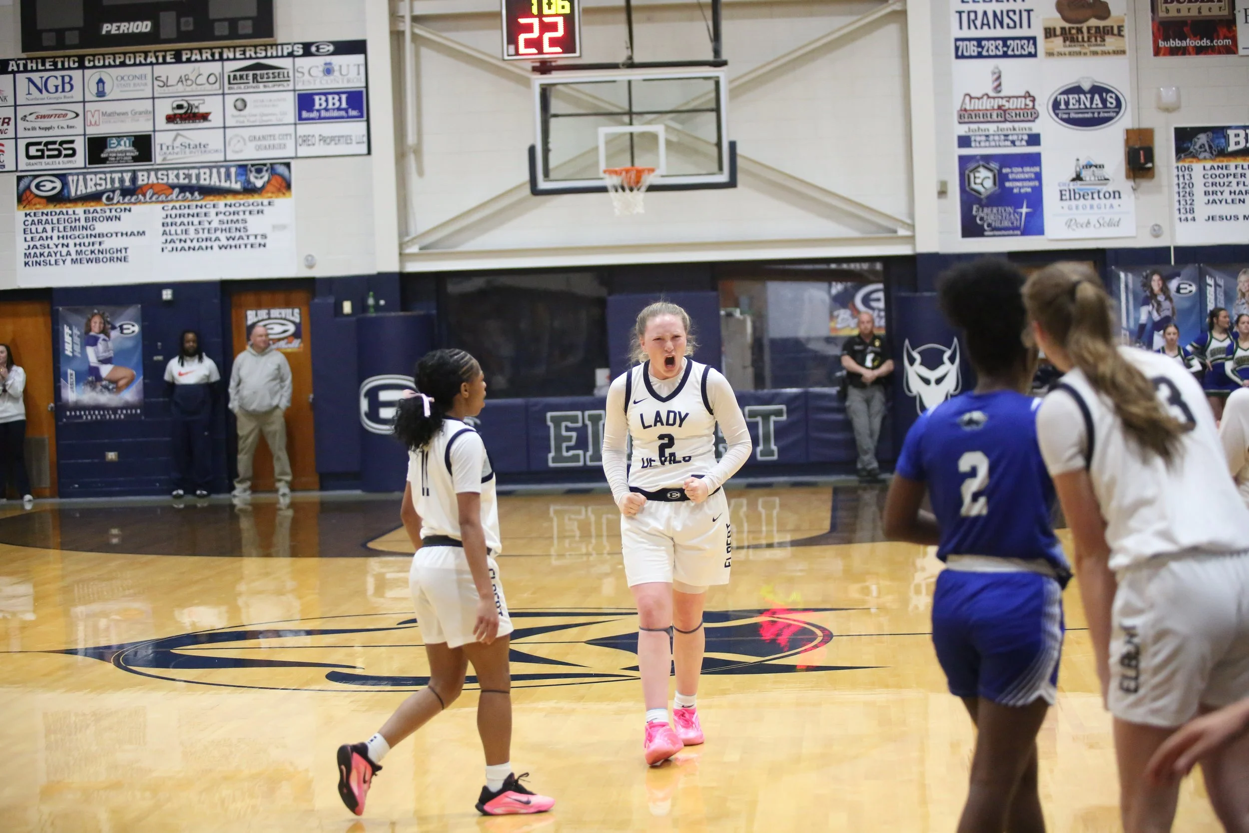 A group of young female basketball players in uniforms on a basketball court, with one player in the center expressing excitement or shouting, and other players facing away or toward her. The gym has banners and advertisements on the walls, a basketb