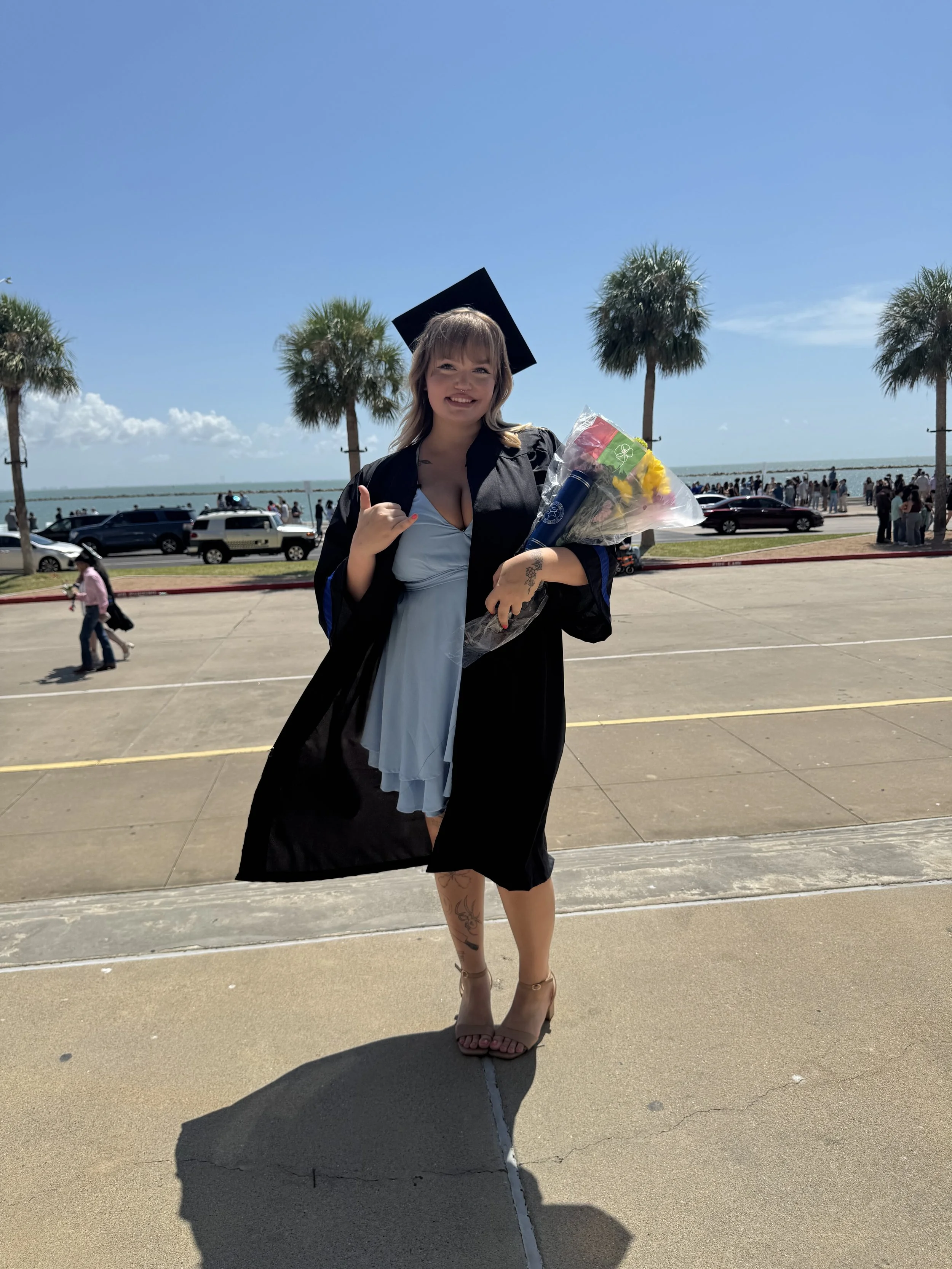 Owner of KM Marketing Management in a graduation cap and gown standing outdoors on a sunny day, holding a bouquet of flowers and celebrating her graduation by giving her universities hand sign, a shakas-up.