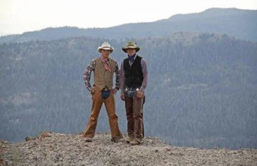 Two men dressed in vintage cowboy attire standing outdoors on rocky terrain with mountains in the background.