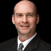 A man in a suit and tie smiling at the camera, with a plain black background.