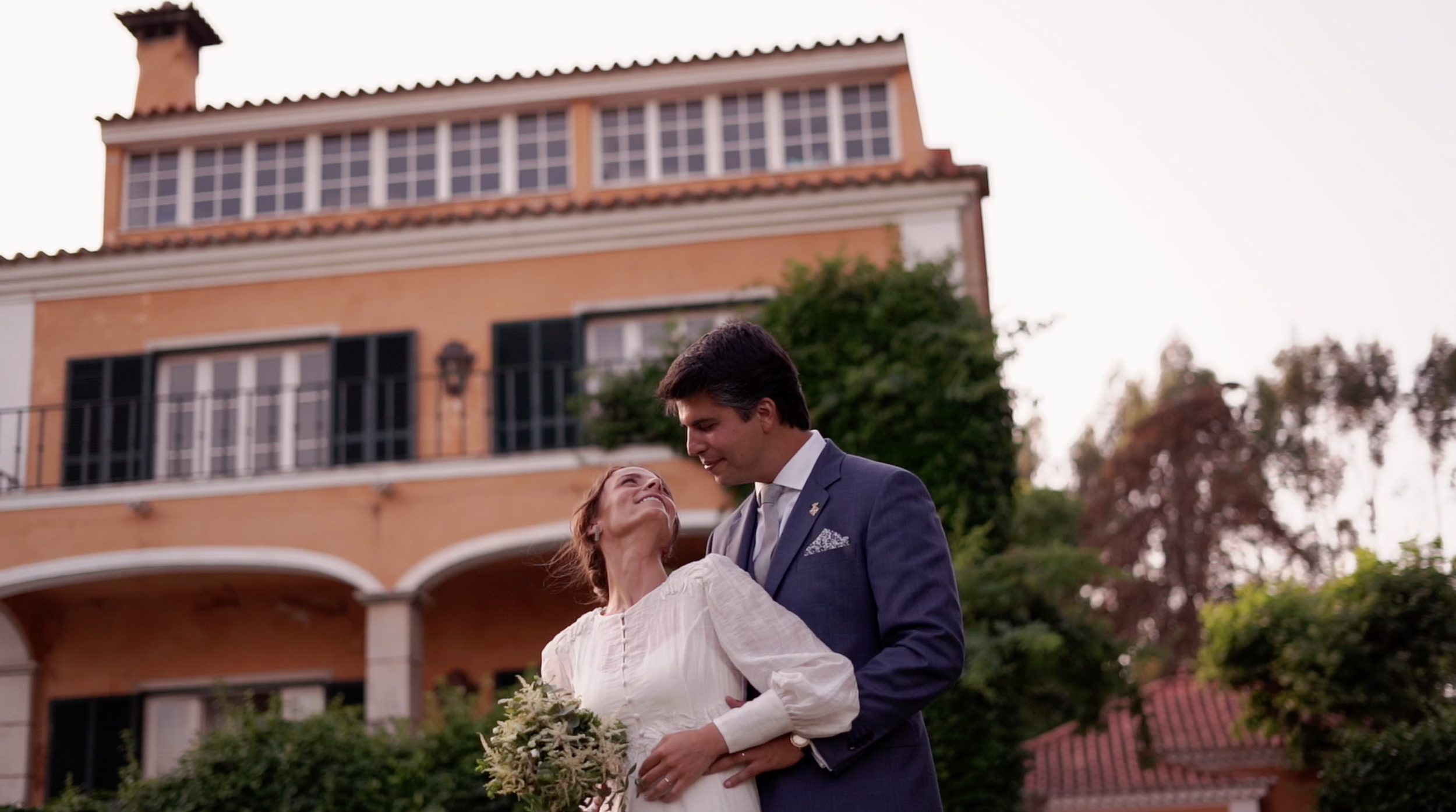 A couple dance in front of a large, orange house with multiple windows and a rooftop. The woman holds a bouquet and looks up at the man, who is smiling at her while dressed in a blue suit.