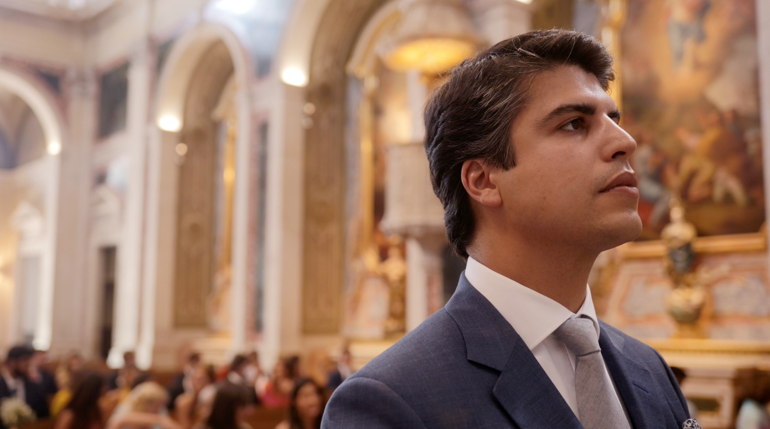 A young man in a suit attending a formal event or ceremony in an ornate, gold-decorated hall with arches and chandeliers.