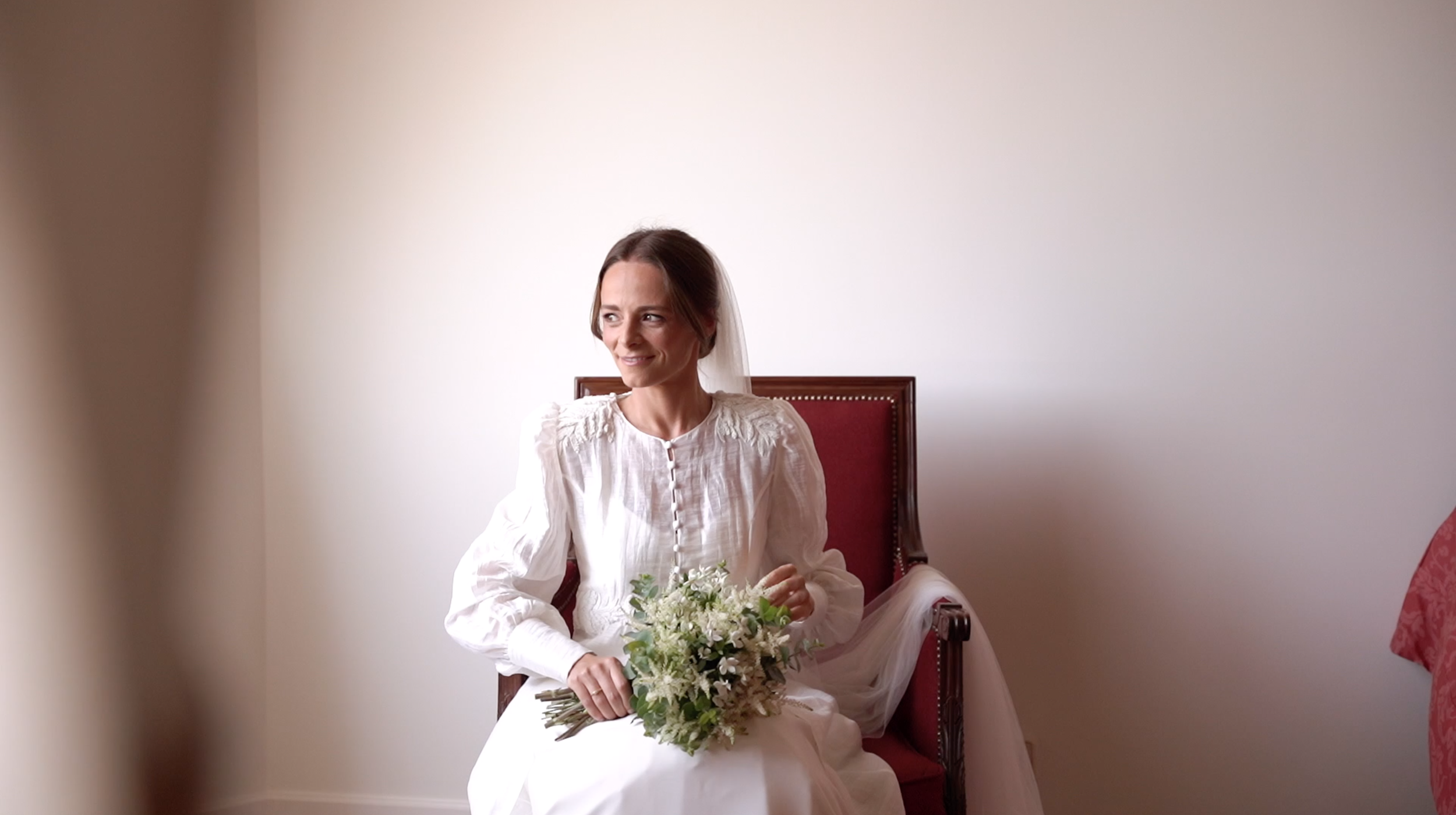 A woman in a white dress with a veil, holding a small bouquet, sitting on a red-upholstered chair against a plain white wall, smiling softly.